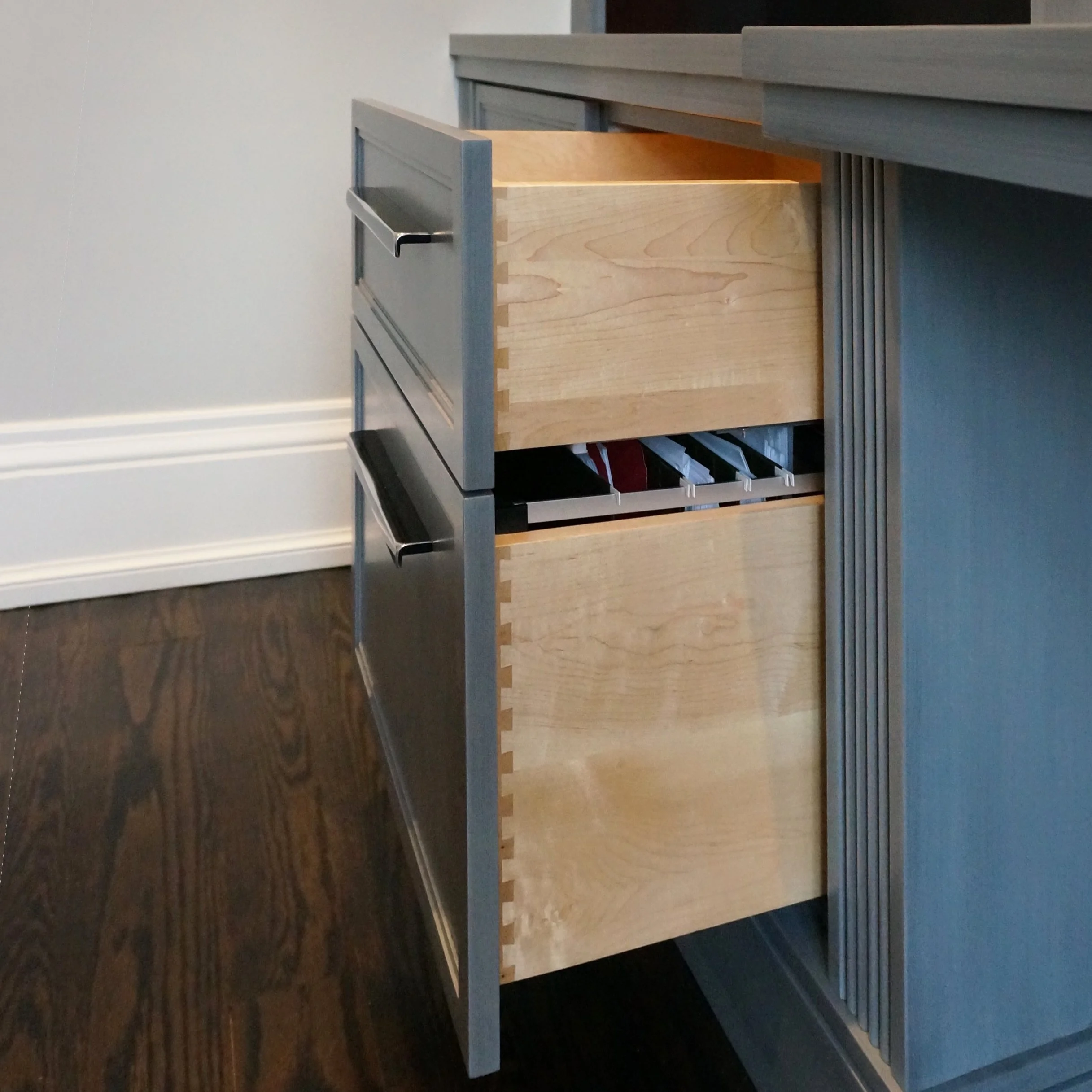 A partially opened wooden drawer protrudes from a gray cabinet, situated on a dark wooden floor in a room with plain white walls.