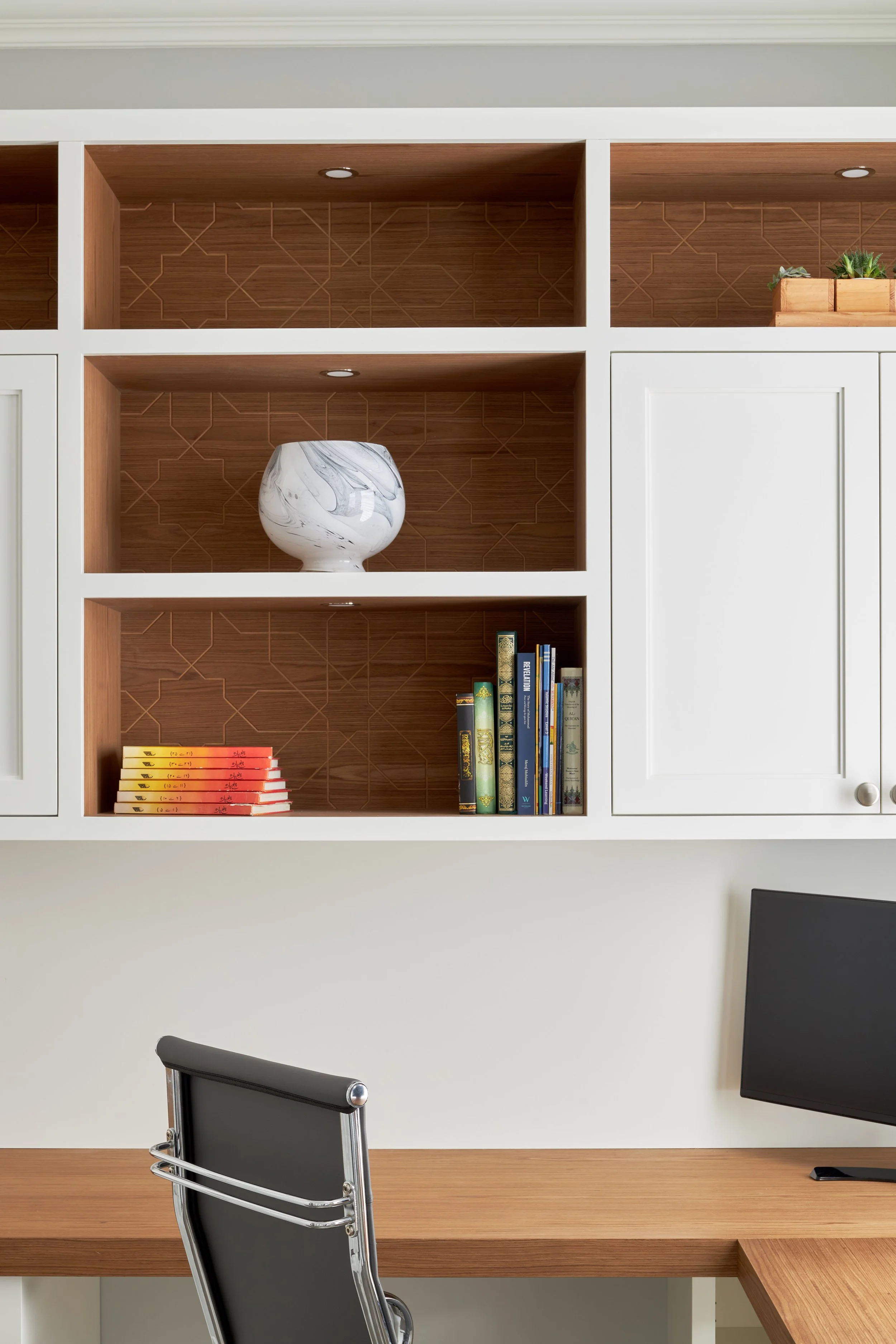 Office desk with a chair, a speaker, a computer monitor, and a wooden cabinet with decorative items and books. Shelves with decorative objects and books are above the desk.