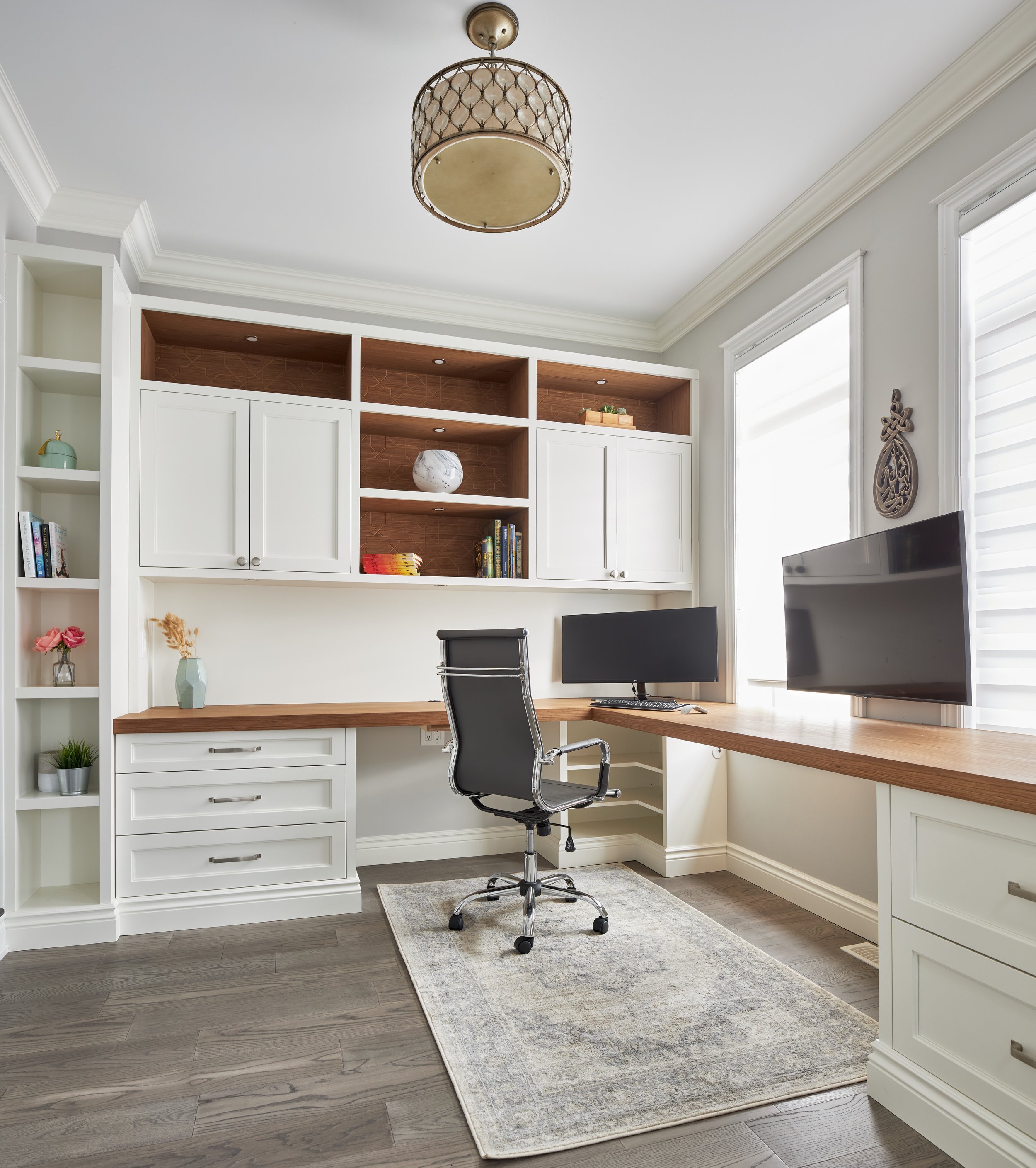 A modern home office with a white built-in bookshelf, a wooden desk with a computer monitor, an office chair, and a gray rug on dark wooden flooring. Natural light comes through window blinds.