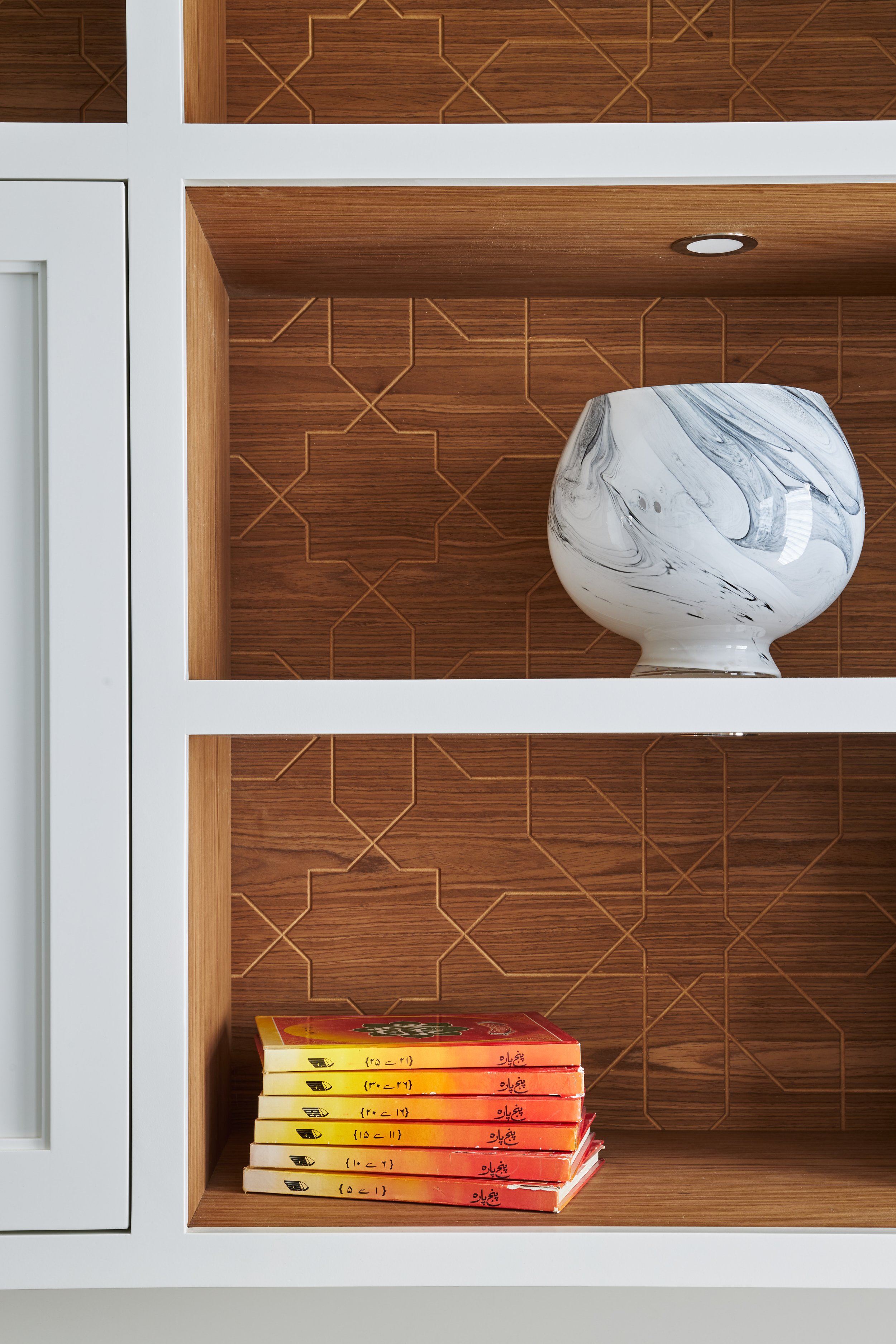 A wooden shelf with a white marble-patterned vase on the right and a stack of seven books with colorful covers on the left. The background features a patterned wooden wall and a white frame surrounding the shelf.