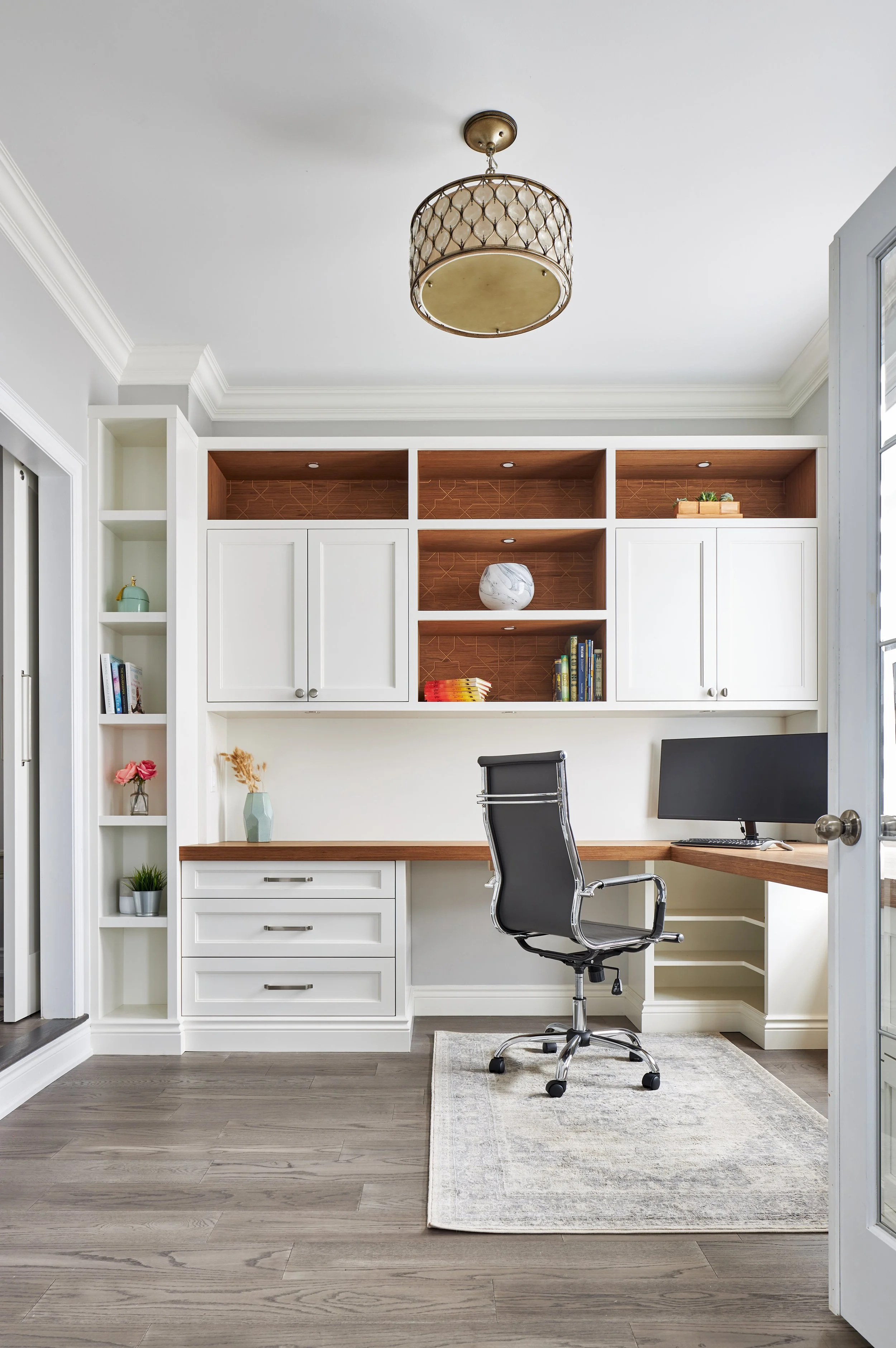 Home office with white built-in shelves, a wooden desk, and an office chair, decorated with plants and books, with a gray rug and wood flooring.