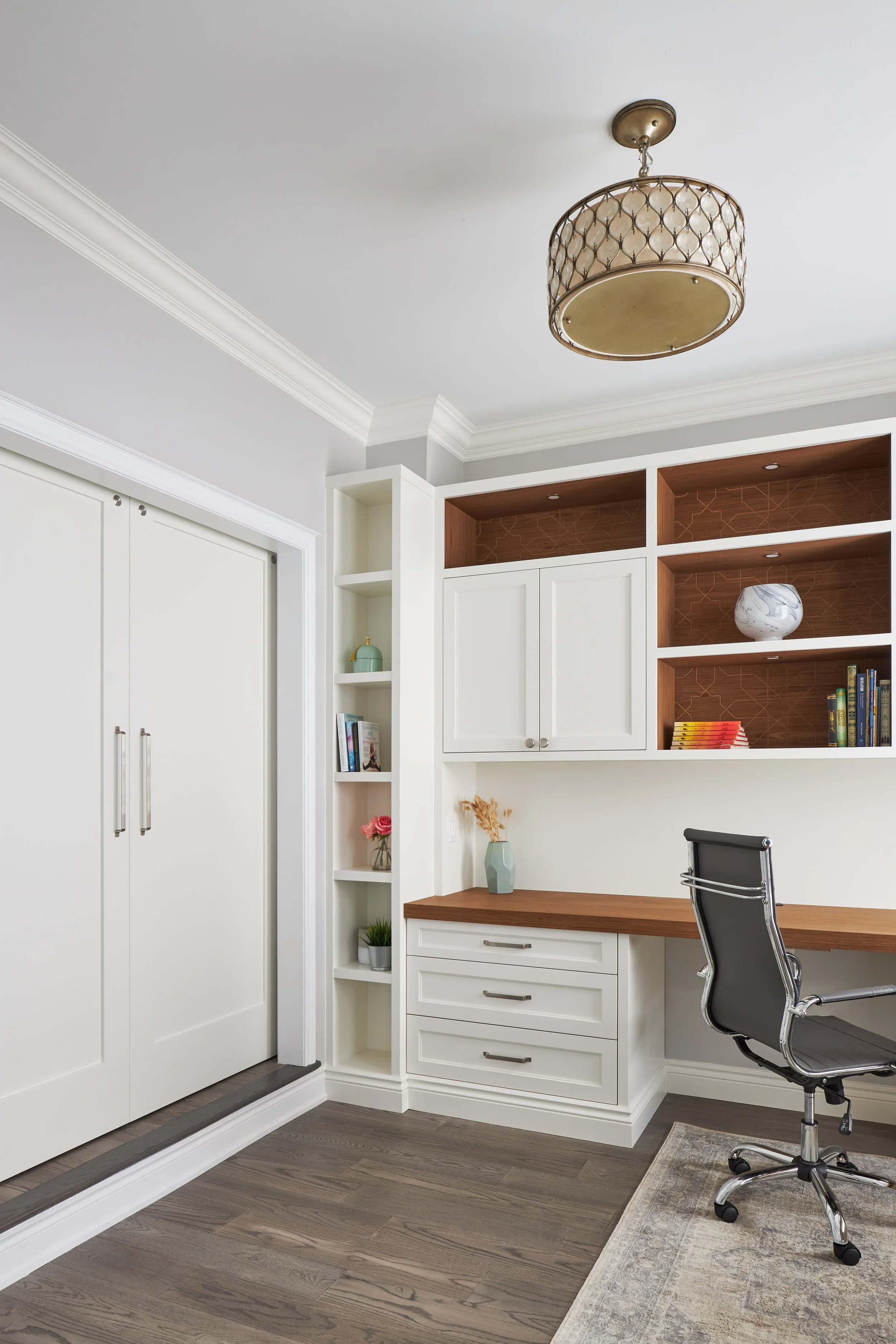 Home office with white built-in shelves, a wooden desk, a black office chair, and decorative items including vases and books. Wooden flooring and a white ceiling with crown molding.