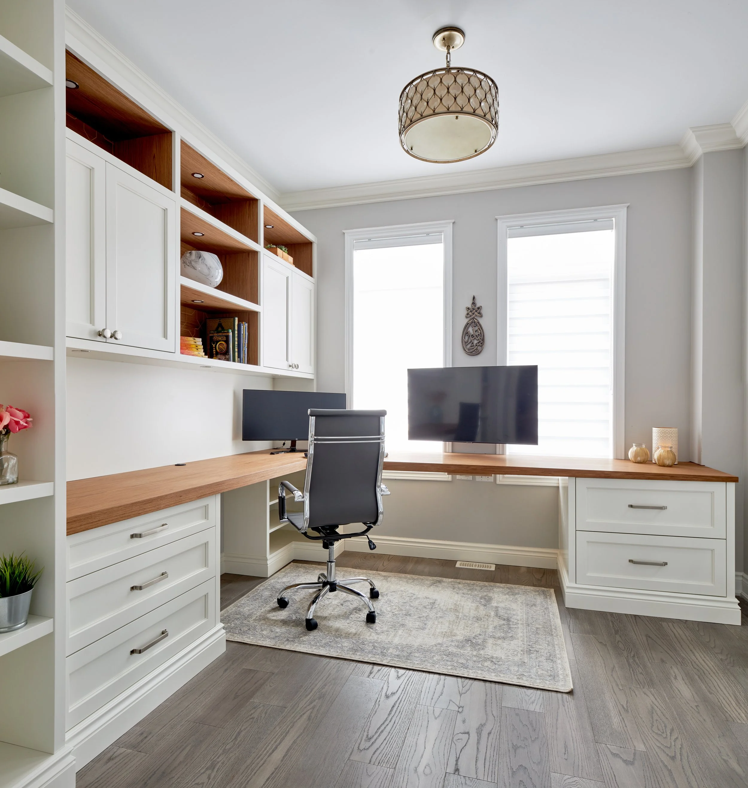 A home office with a white built-in bookshelf, a wooden desk, and a black office chair in front of two large windows. The room has light gray walls, white trim, hardwood floors, and decorative ceiling molding. There are three decorative vases on a sm