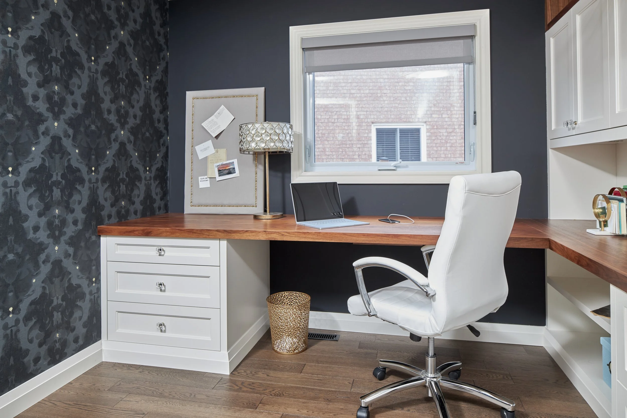 A home office with a wooden desk, white swivel chair, and dark blue wall decorated with gold star-like accents and black patterned wallpaper on an adjacent wall. A window with a gray roller shade provides natural light. There is a white built-in cabi