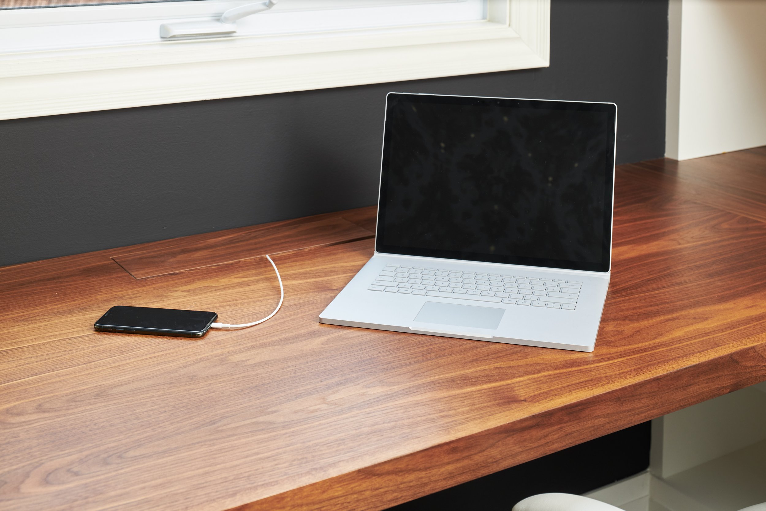 A silver laptop with a black screen on a wooden desk, connected to a black smartphone via a white charging cable, with a window in the background.