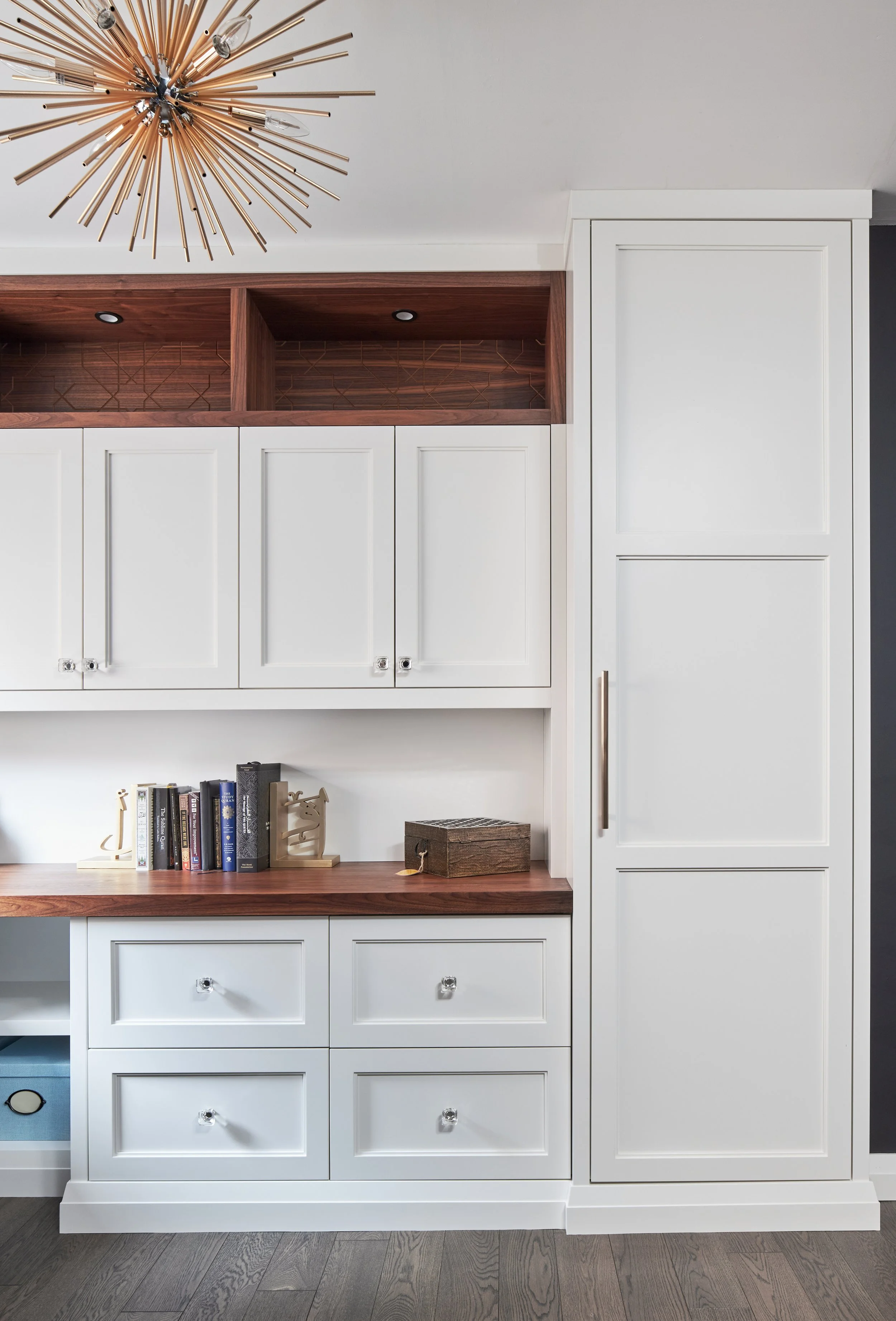 A white built-in cabinet with drawers and closed doors, wooden countertop, open shelves with wood finish, books, decorative items, a wooden box, and a chandelier with gold-colored rods on the ceiling.