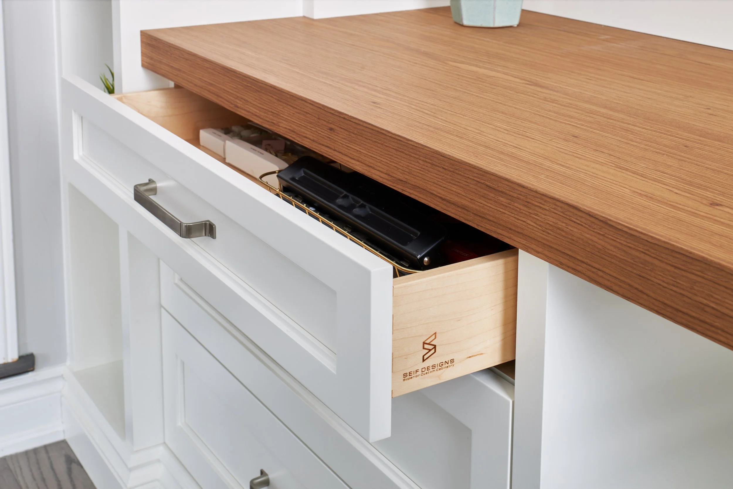A white cabinet with a sliding wooden countertop partially slid out, revealing storage underneath containing a black item, a cardboard box, and some small objects.