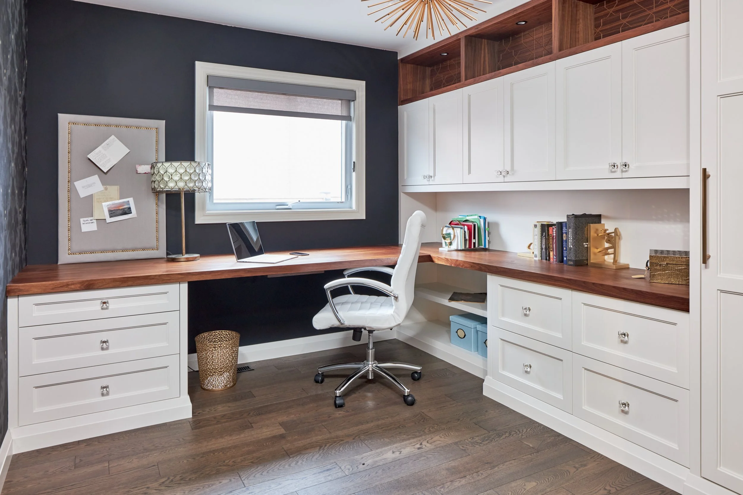 Home office with a built-in white desk and cabinets, a window with a roller shade, dark gray accent wall, desk lamp, laptop, books, and a white office chair.