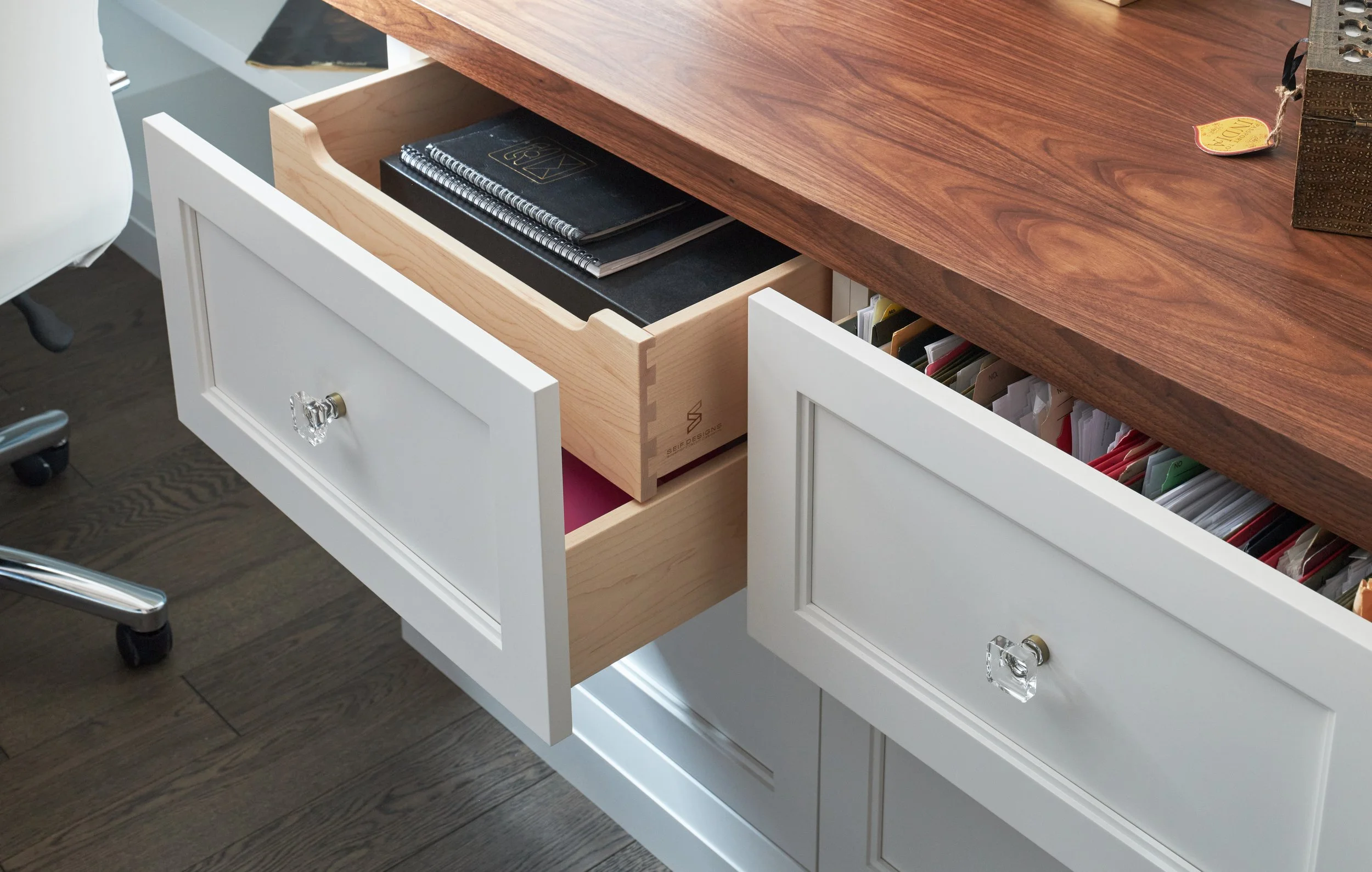 A white desk with two open drawers; the top drawer contains black notebooks and spiral notebooks, and the bottom drawer has hanging file folders. Part of an office chair with wheels is visible to the left.