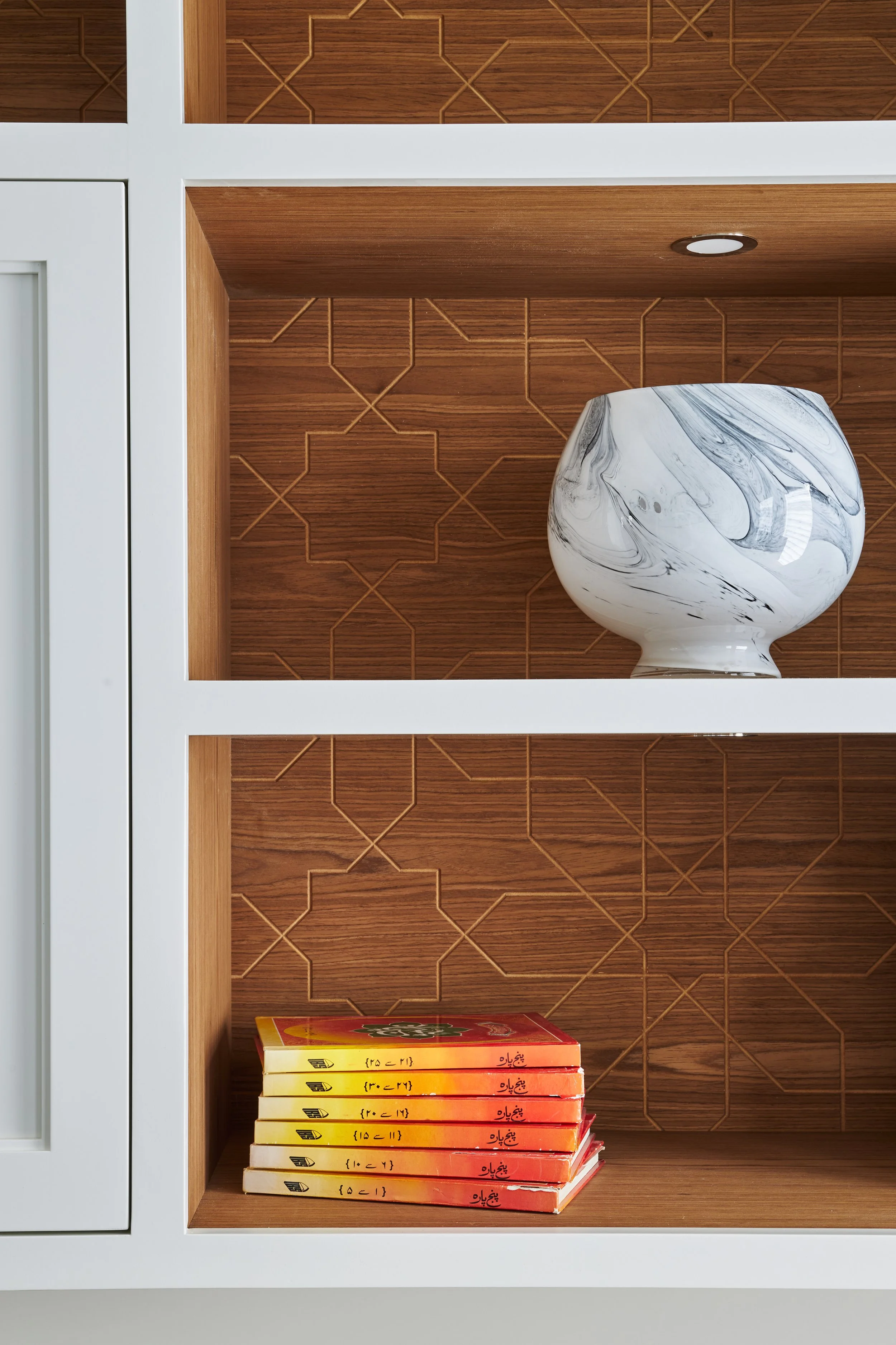 A bookshelf with a marble-patterned vase on the middle shelf and a stack of books with yellow, orange, and red covers on the bottom shelf. The background features brown patterned wooden panels and a small recessed ceiling light.