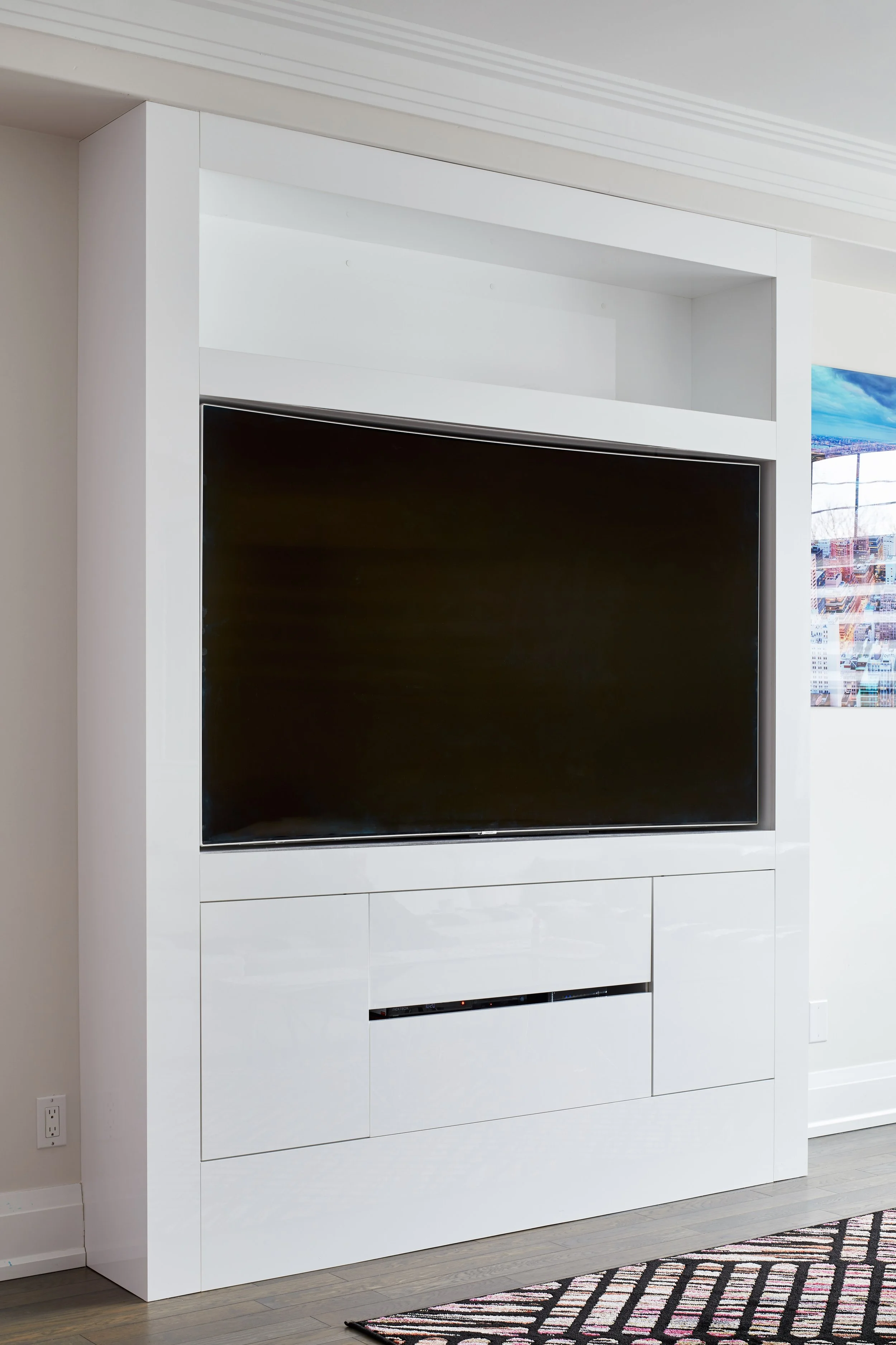 A large flat screen television installed within a white built-in cabinet with closed drawers below. The cabinet is within a modern living room with a striped rug on light hardwood flooring, and a cityscape view visible through the window on the right