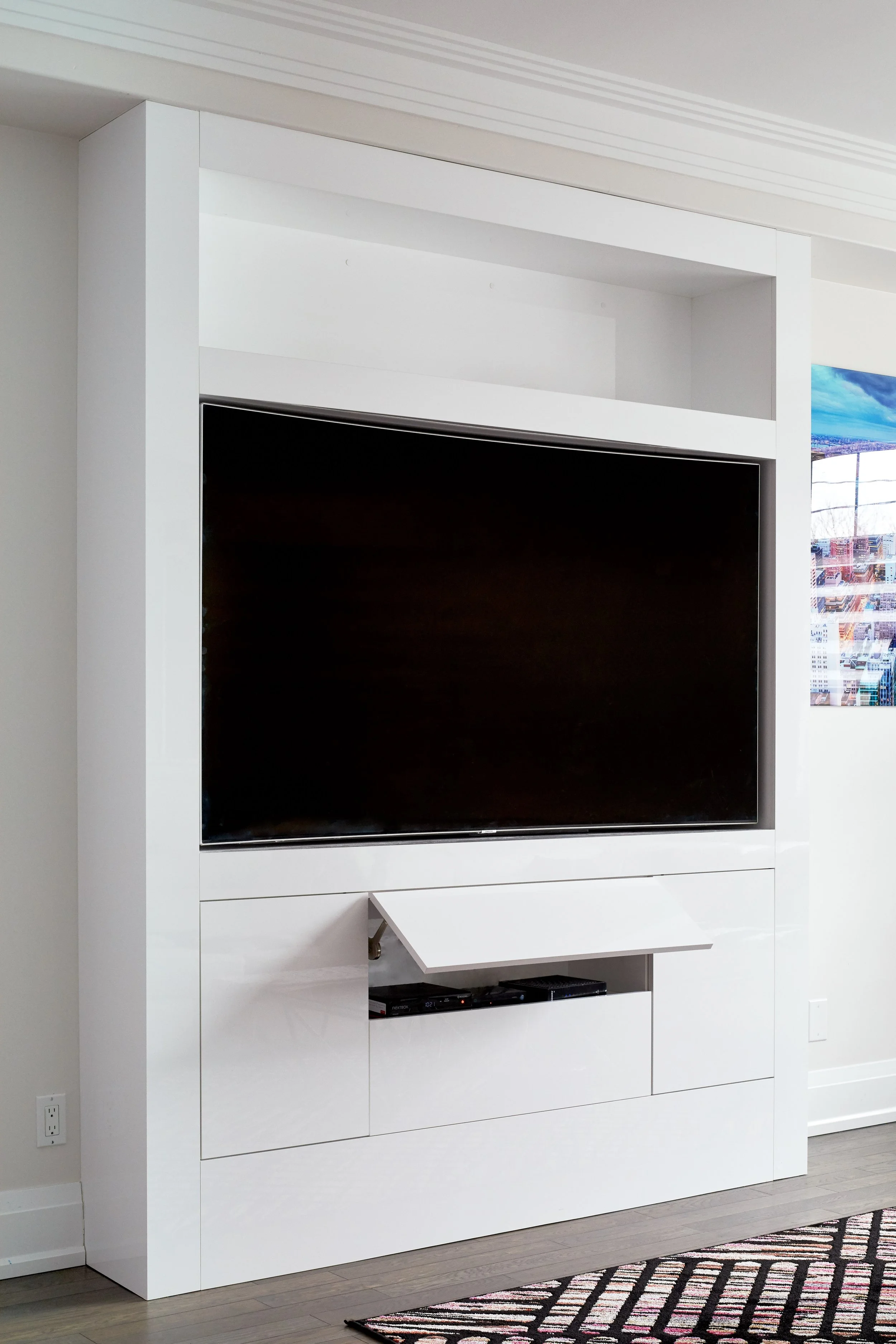 A modern white entertainment center with a flat-screen television and electronic devices, in a room with light-colored hardwood floors and a patterned rug.