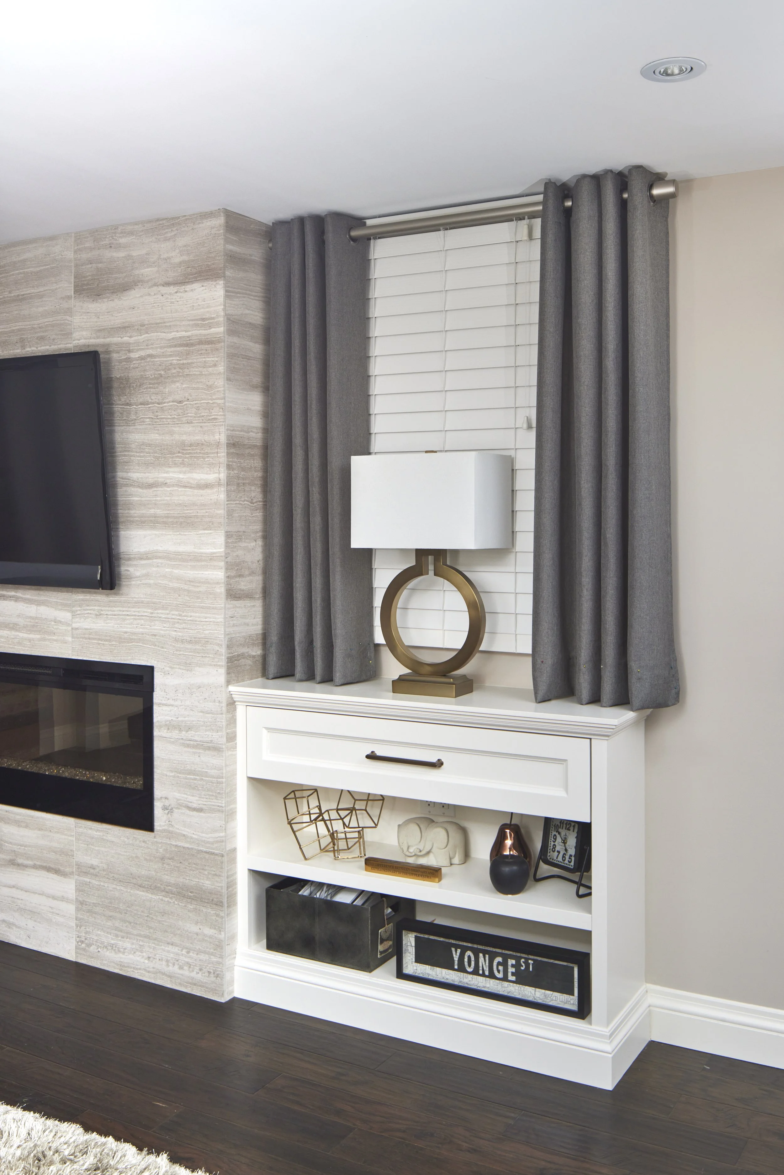 Interior living space with a fireplace, beige marble accent wall, and a white bookshelf decorated with various objects, including a black vase, clock, and metal sculptures. Grey curtains hang in front of a window with white blinds.