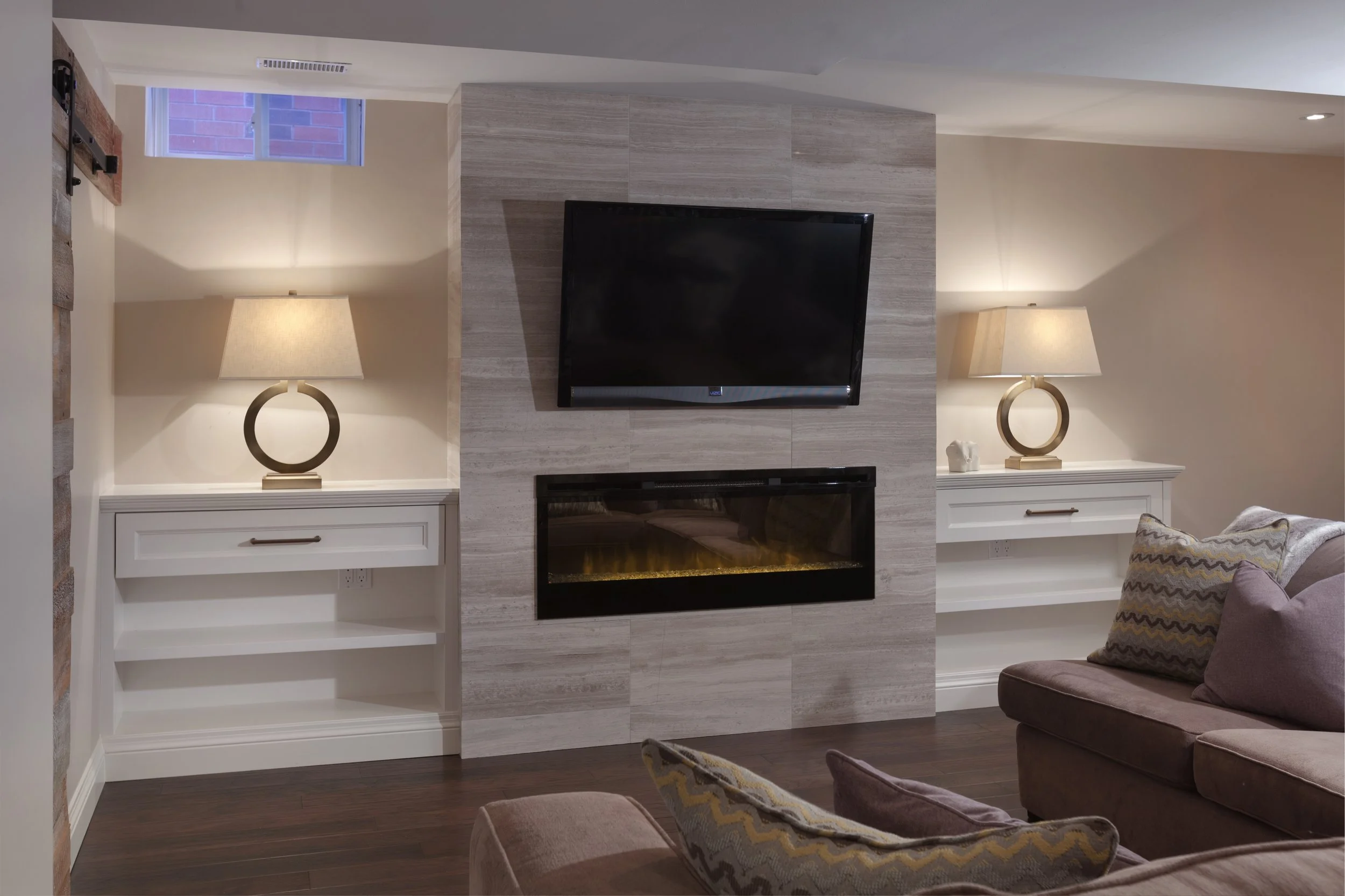 Living room featuring a flat-screen TV mounted on a stone accent wall, two matching table lamps on white side tables, a fireplace beneath the TV, a couch with patterned pillows, and a window with brick view.