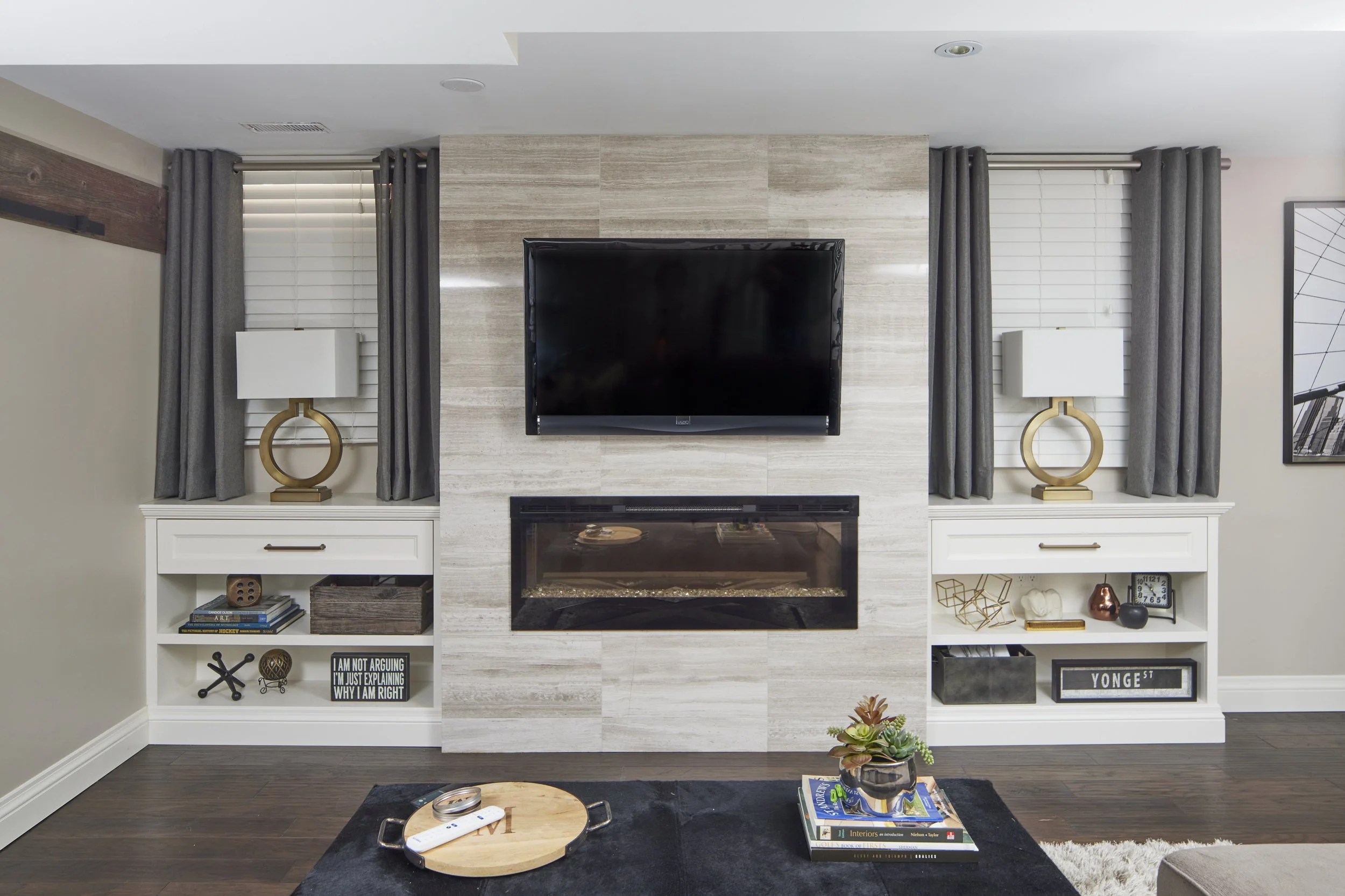 Living room with fireplace and built-in white shelves decorated with books, art, and ornaments. Two side tables with lamps, a flat-screen TV above the fireplace, curtains on windows, and a small plant on the floor.
