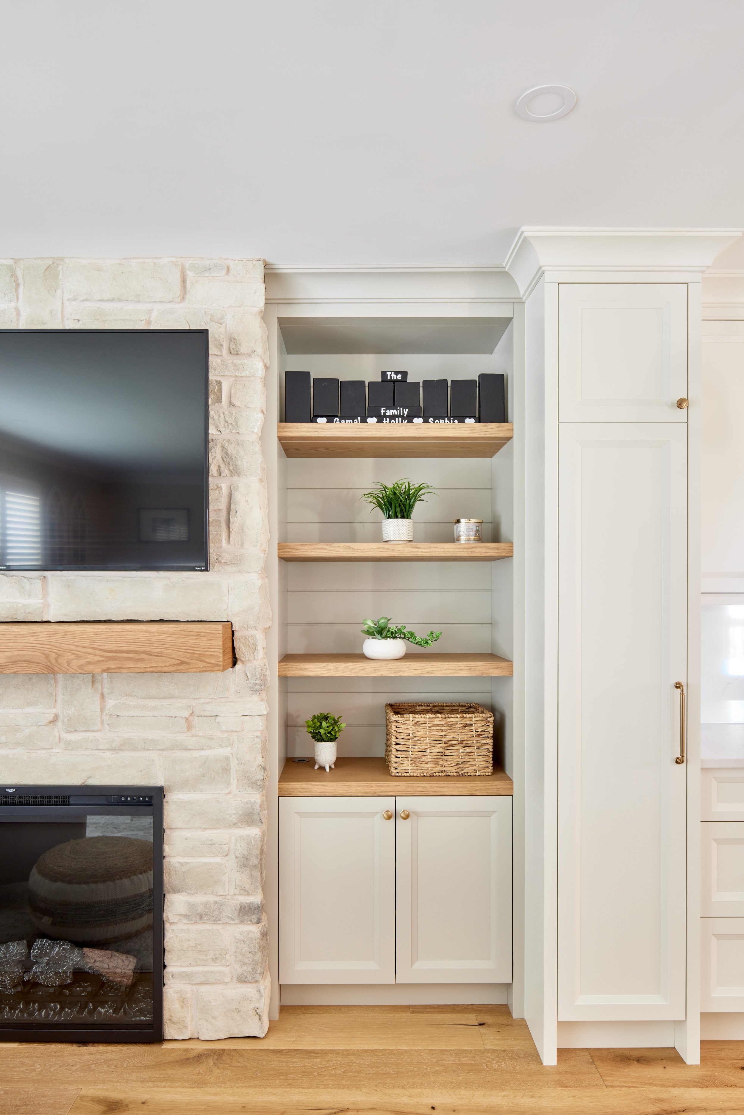 Living room with a flat-screen TV mounted on a stone wall, and a white built-in bookcase with shelves containing plants, a wicker basket, and decorations, next to a white cabinet with gold handles.