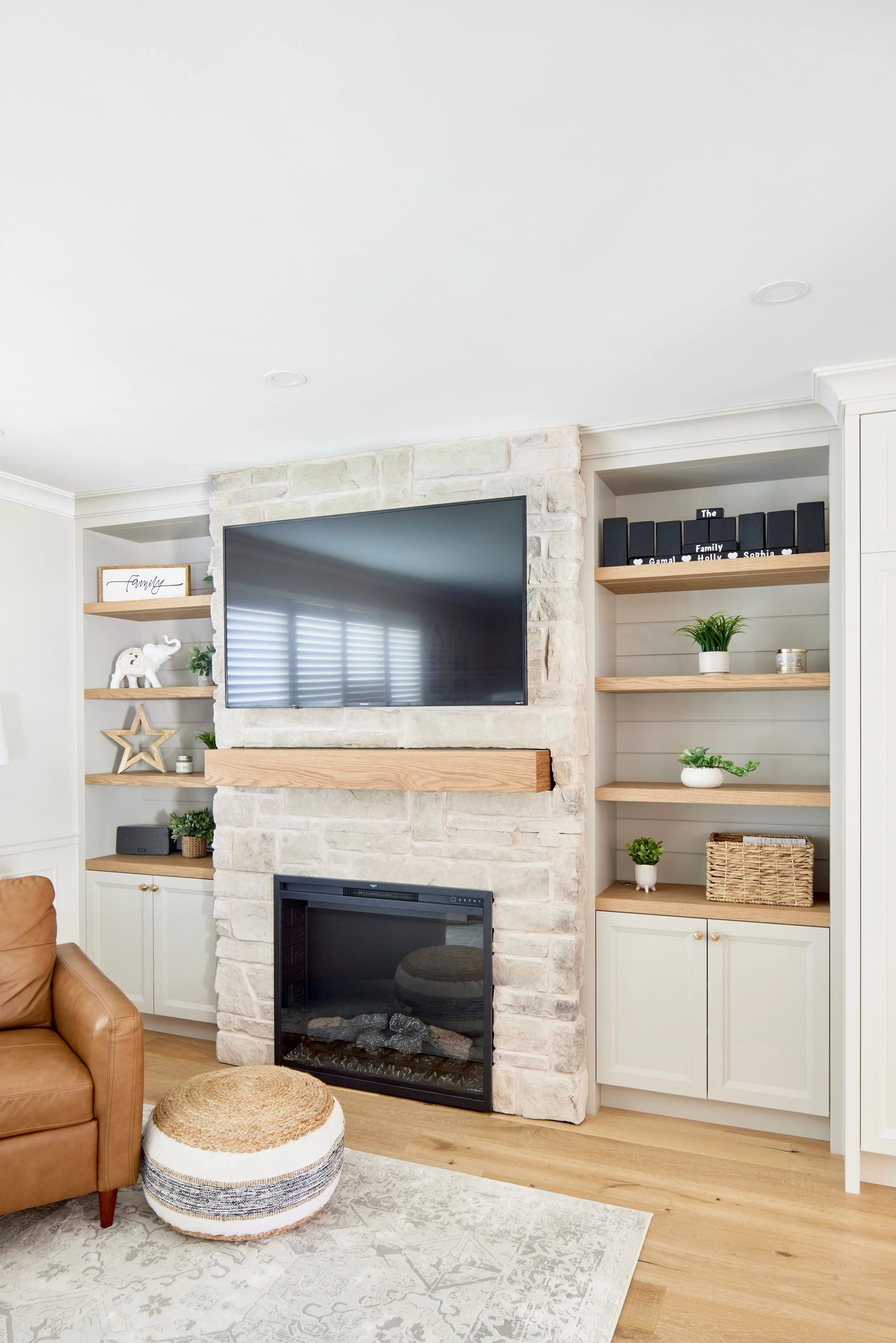 Living room with white brick fireplace, mounted TV, built-in shelves with decor, and light wood flooring.
