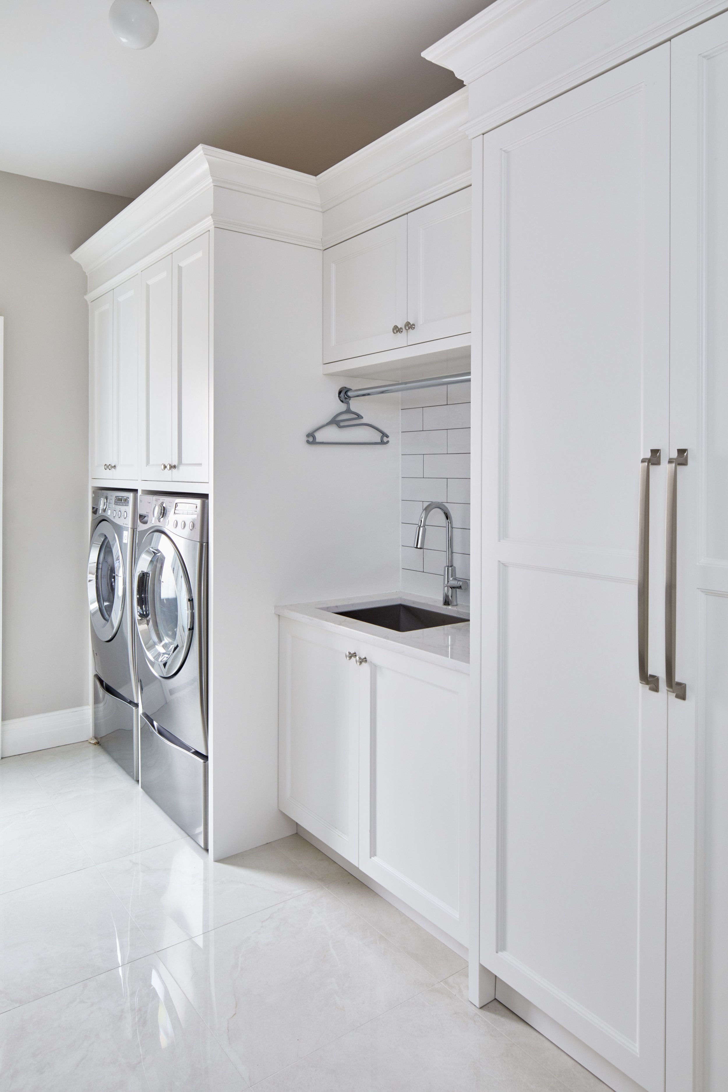 A laundry room with white cabinetry, a stackable front-loading washer and dryer, a small countertop sink, and a hanging rod with a single hanger, all on a shiny tile floor.