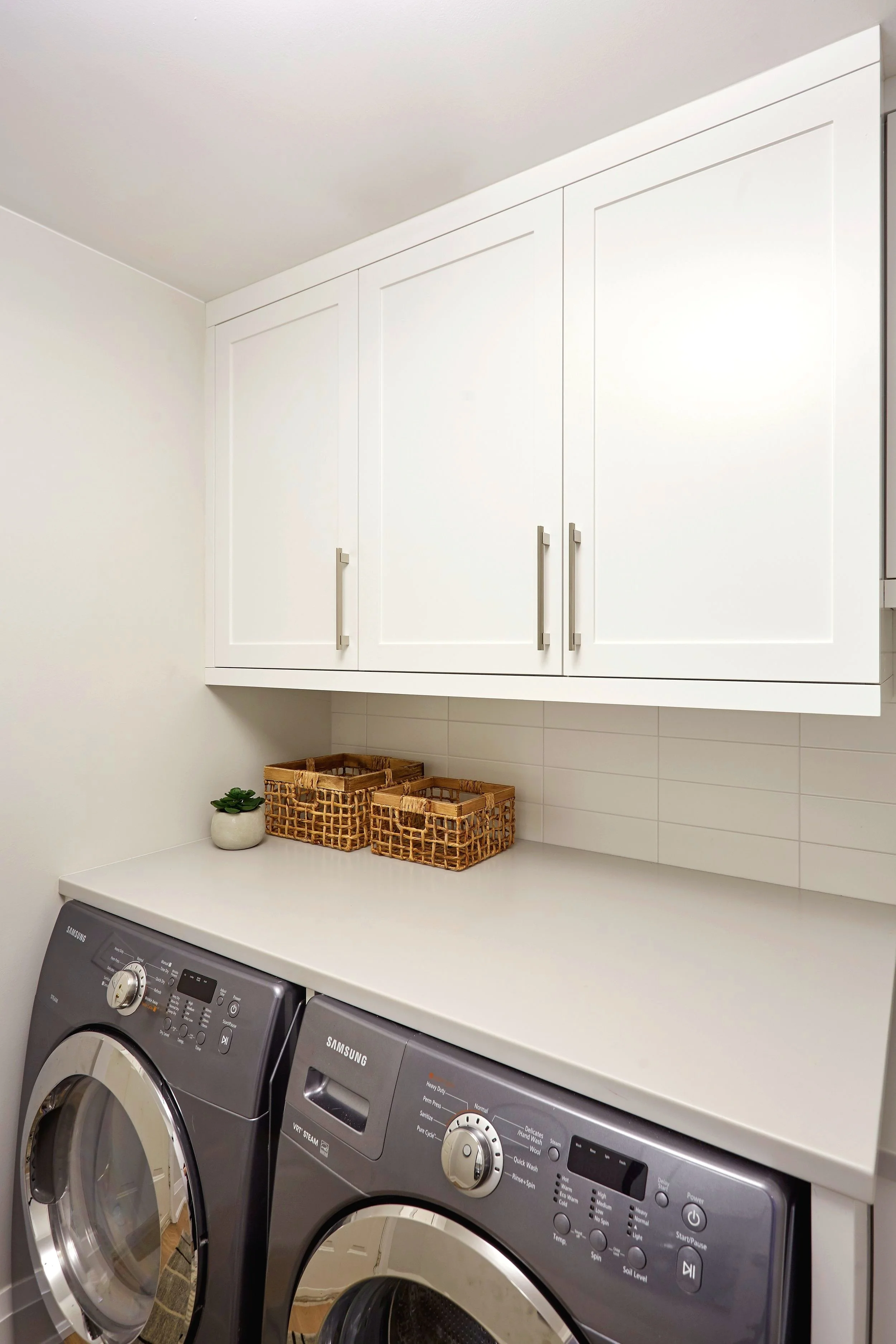 Front load washing machines in a laundry room with a white countertop and white cabinets above. Small baskets and a small plant on the counter.