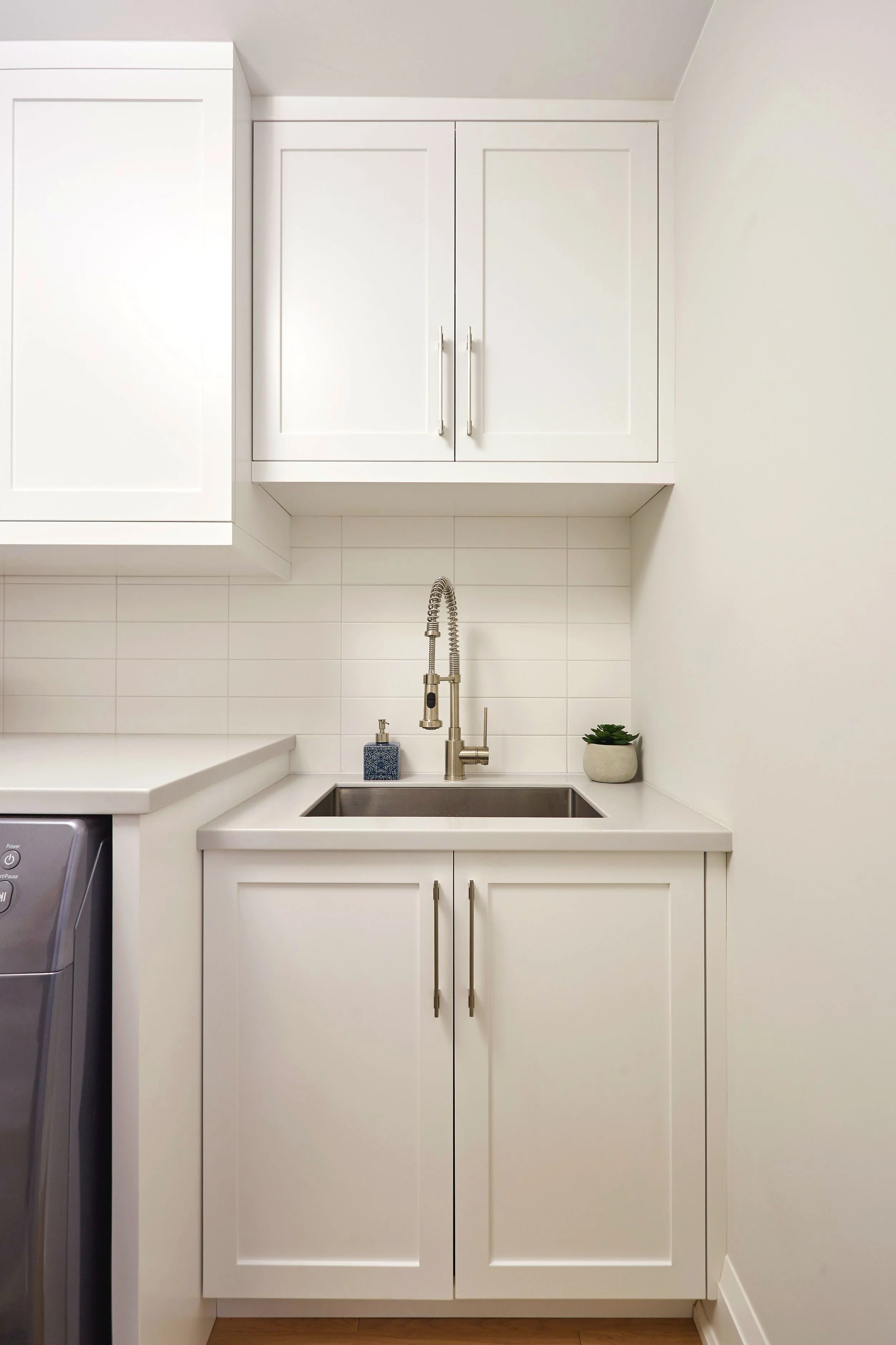 A white kitchen with upper and lower cabinets, a stainless steel sink, and a chrome faucet. There is a small potted plant and a blue soap dispenser on the countertop near the sink. The wall behind the sink has white tiles, and a part of a washing mac