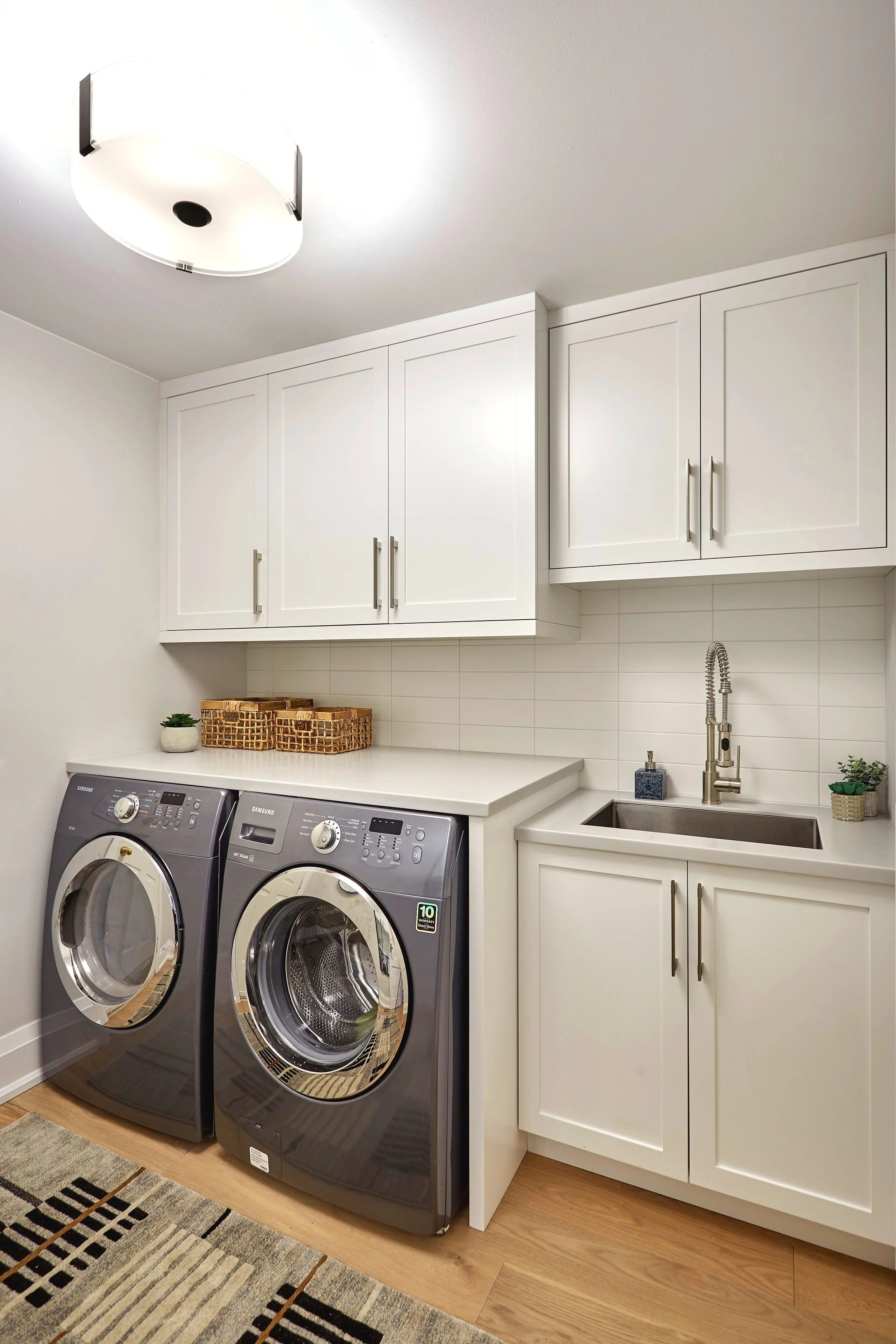 A laundry room with industrial white cabinets, a white countertop, Samsung front-loading washer and dryer in gray, a small potted plant, and woven baskets on the counter.