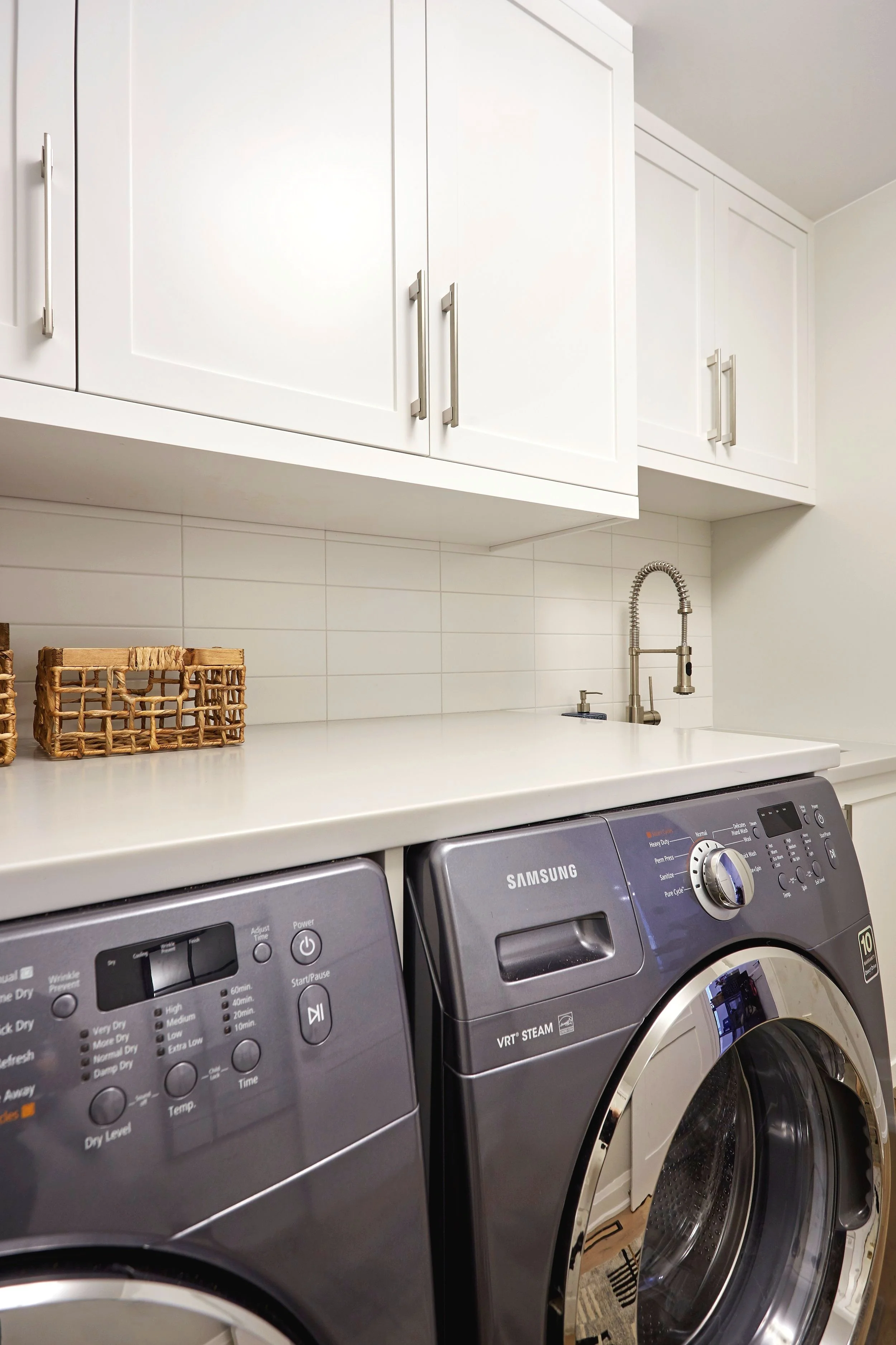 A laundry room with white cabinets above a white countertop, two basket containers on the countertop, and a front-loading Samsung washing machine and dryer side by side, with a silver faucet and sink above the appliances.