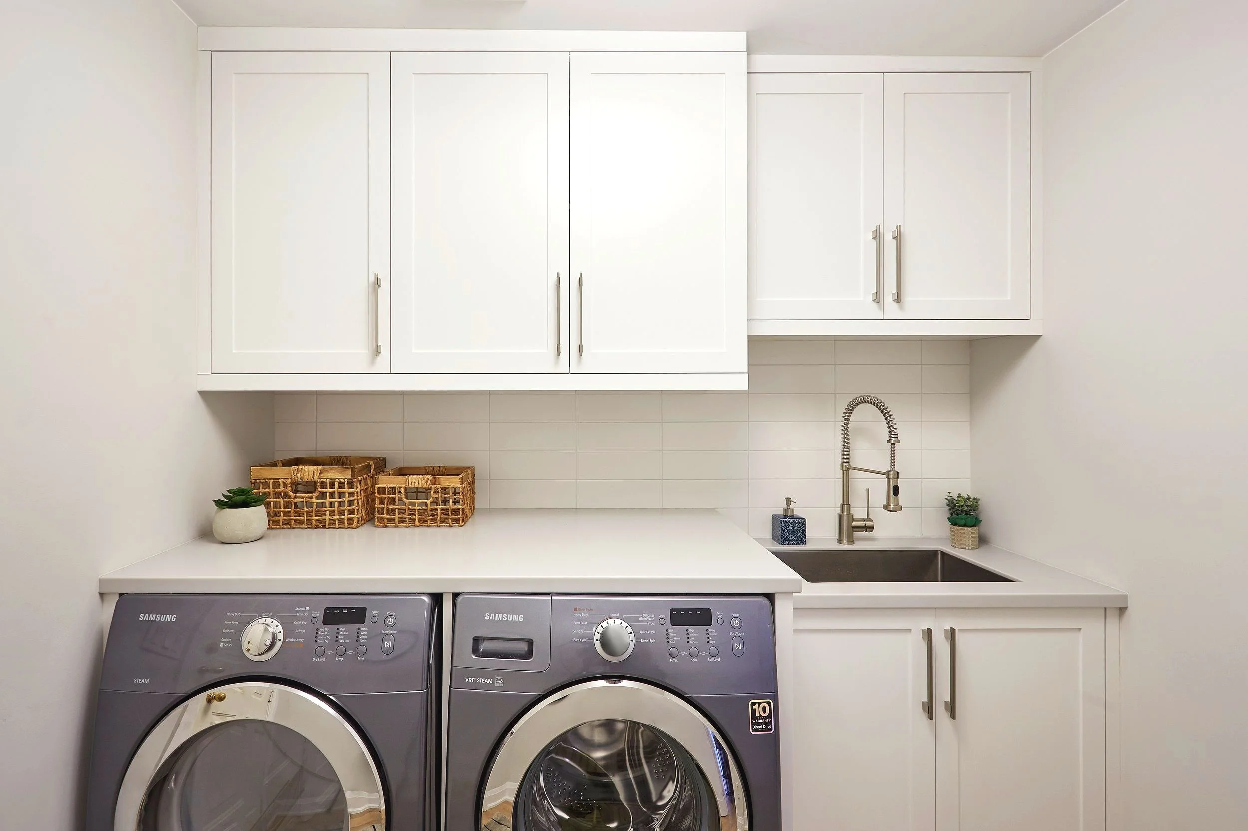A modern laundry room with white cabinets, a countertop, and two washing machines, decorated with small plants and woven baskets.