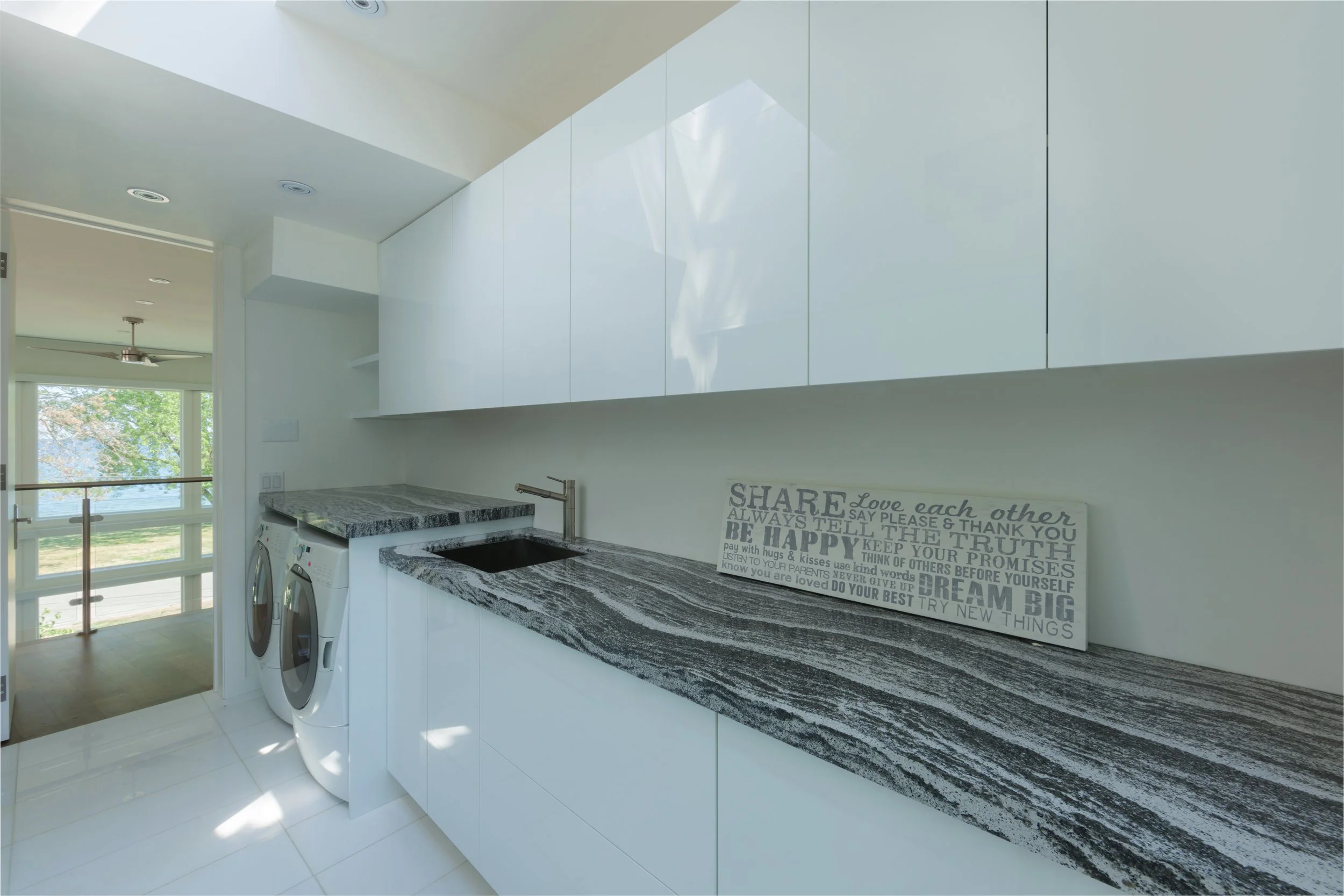 Modern laundry room with white cabinets above a black and white granite countertop, washer and dryer, decorative wall sign with positive words, large window showing green trees outside.