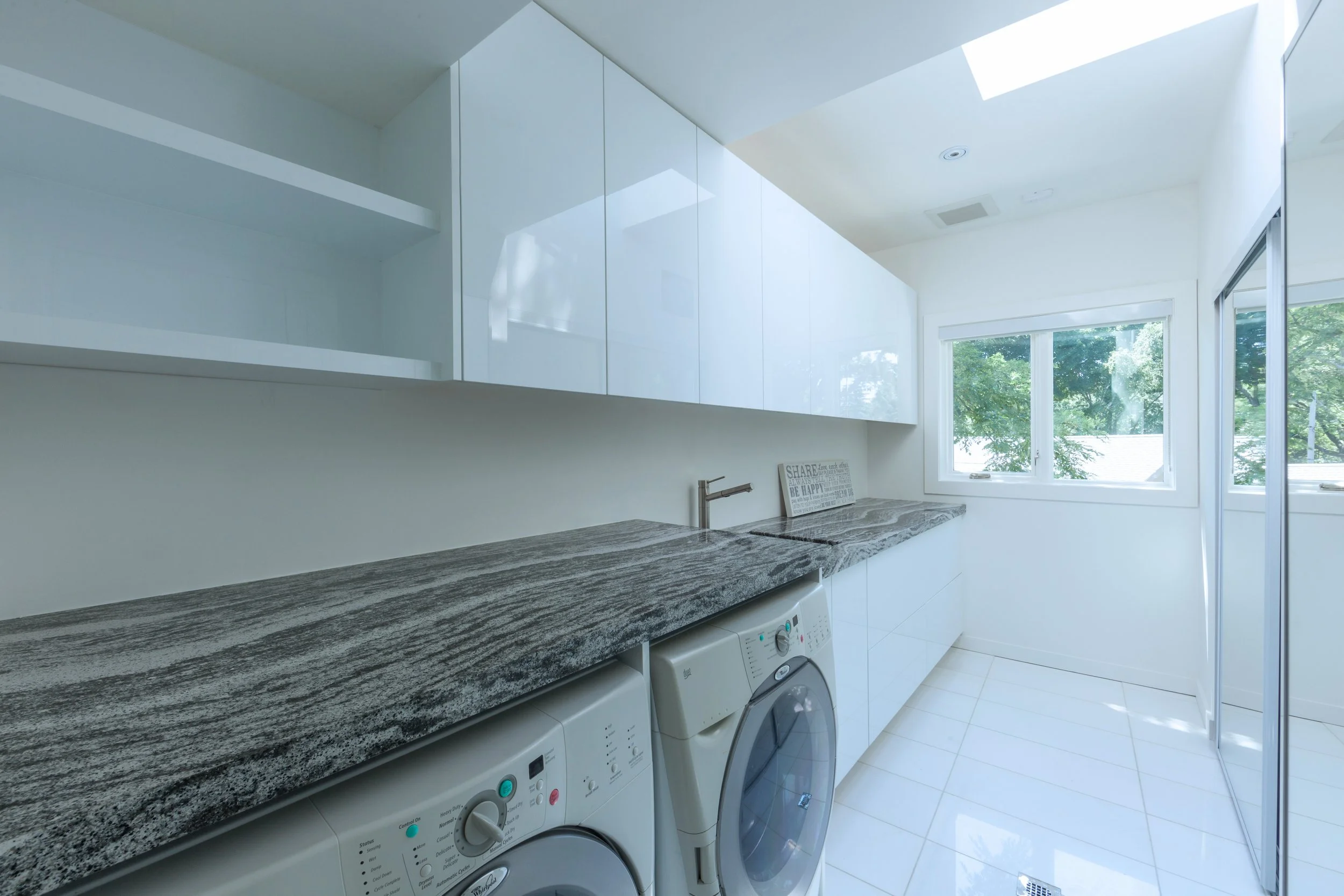 A laundry room with white washing machines underneath a granite countertop, large windows with outside greenery, and white cabinets on the wall.