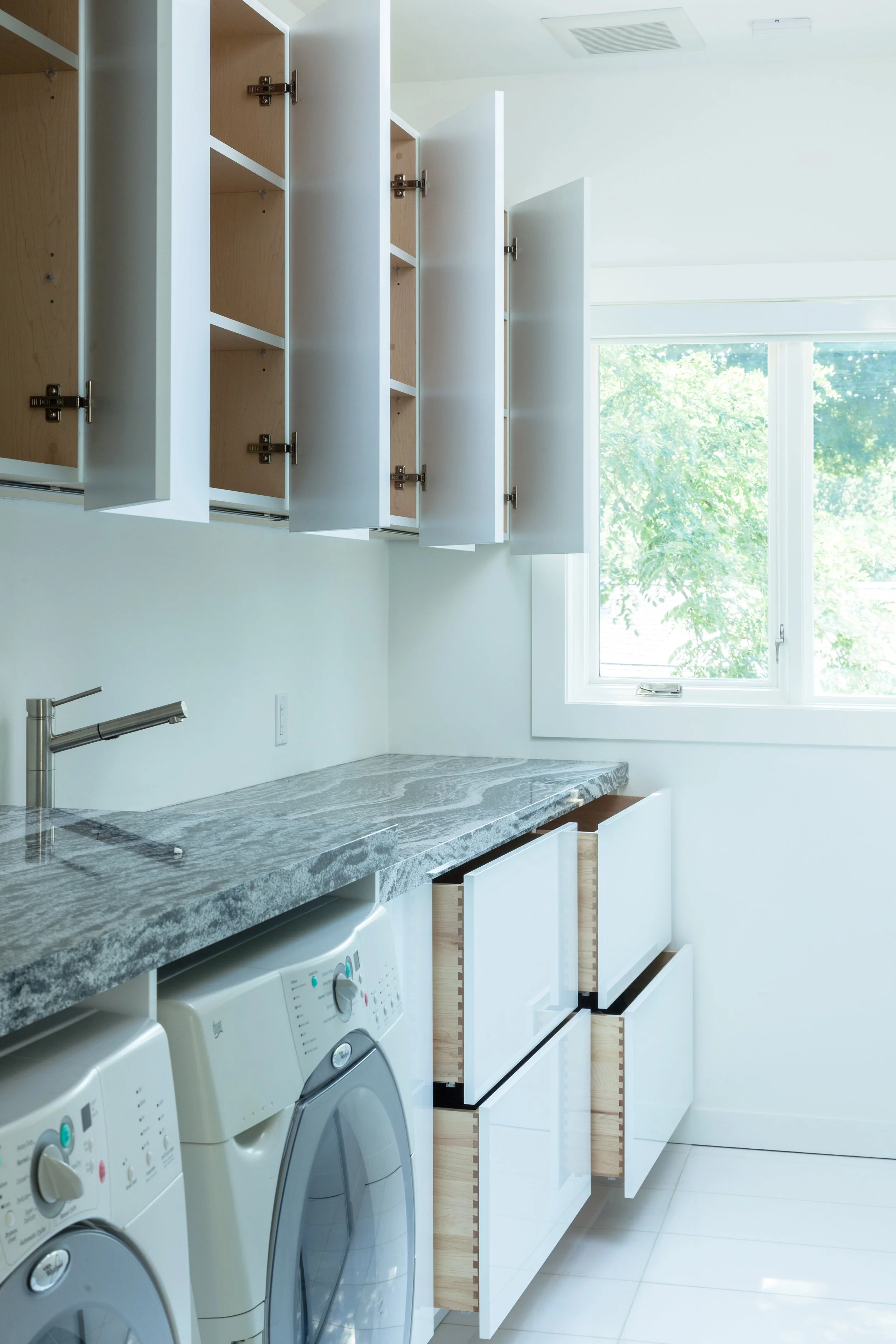 A laundry room with open upper cabinets, a marble countertop, a washing machine, and empty cabinets under the counter, with a large window and outside greenery.