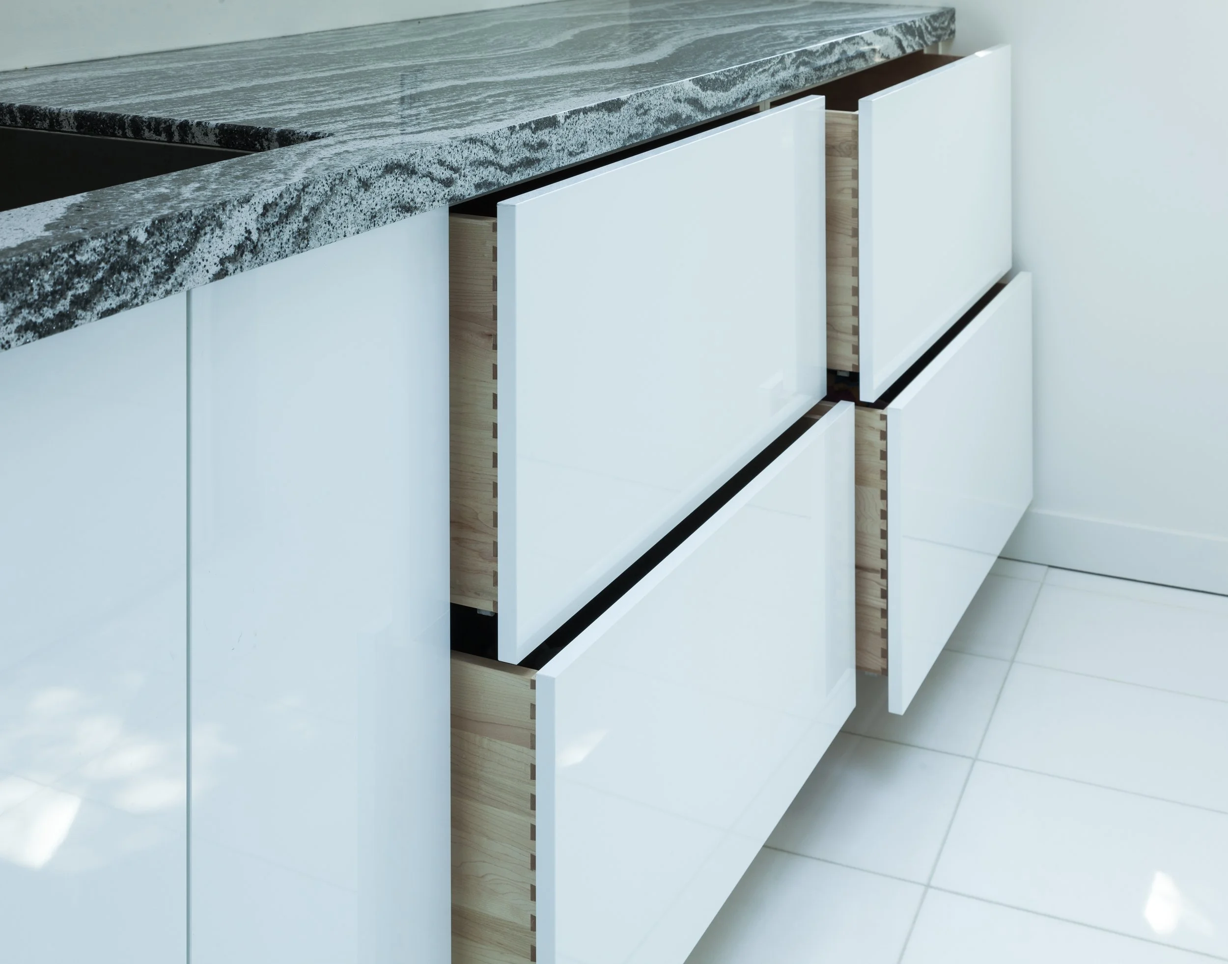 Close-up of white kitchen cabinets with drawers, some partially open, revealing light wood interiors, beneath a gray granite countertop.
