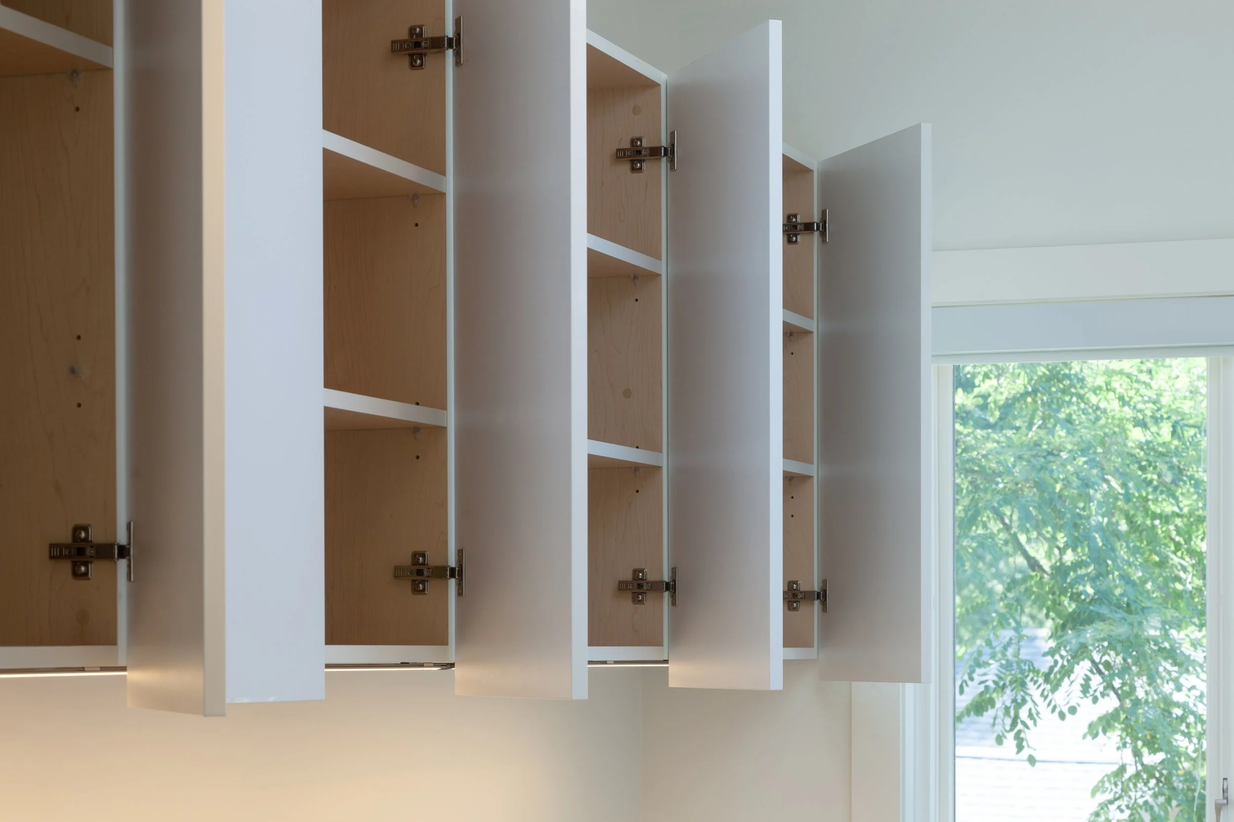 Open white cabinets with wooden shelves, facing a window with green trees outside.