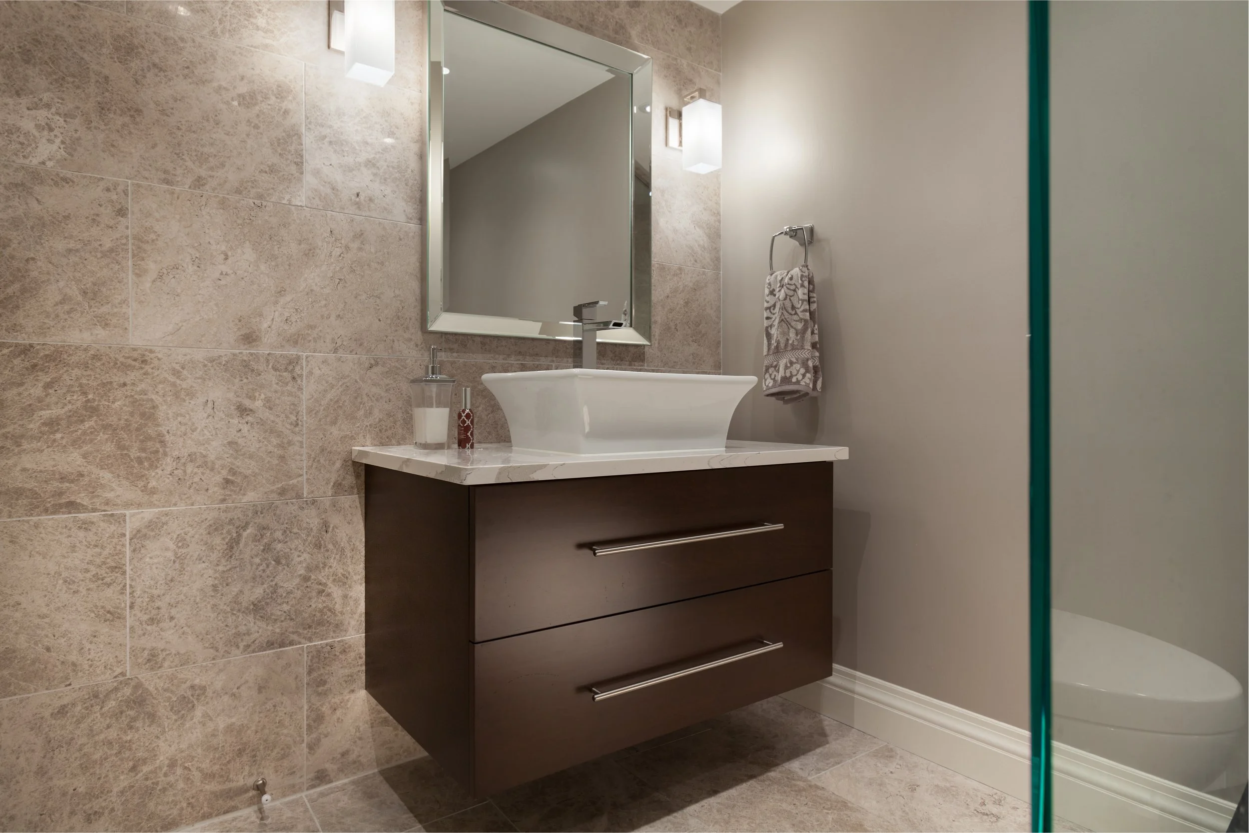 Modern bathroom with beige tiled wall, white sink, and dark wood vanity with two drawers, a mirror, wall-mounted light, towel ring with patterned towel, and glass shower enclosure.