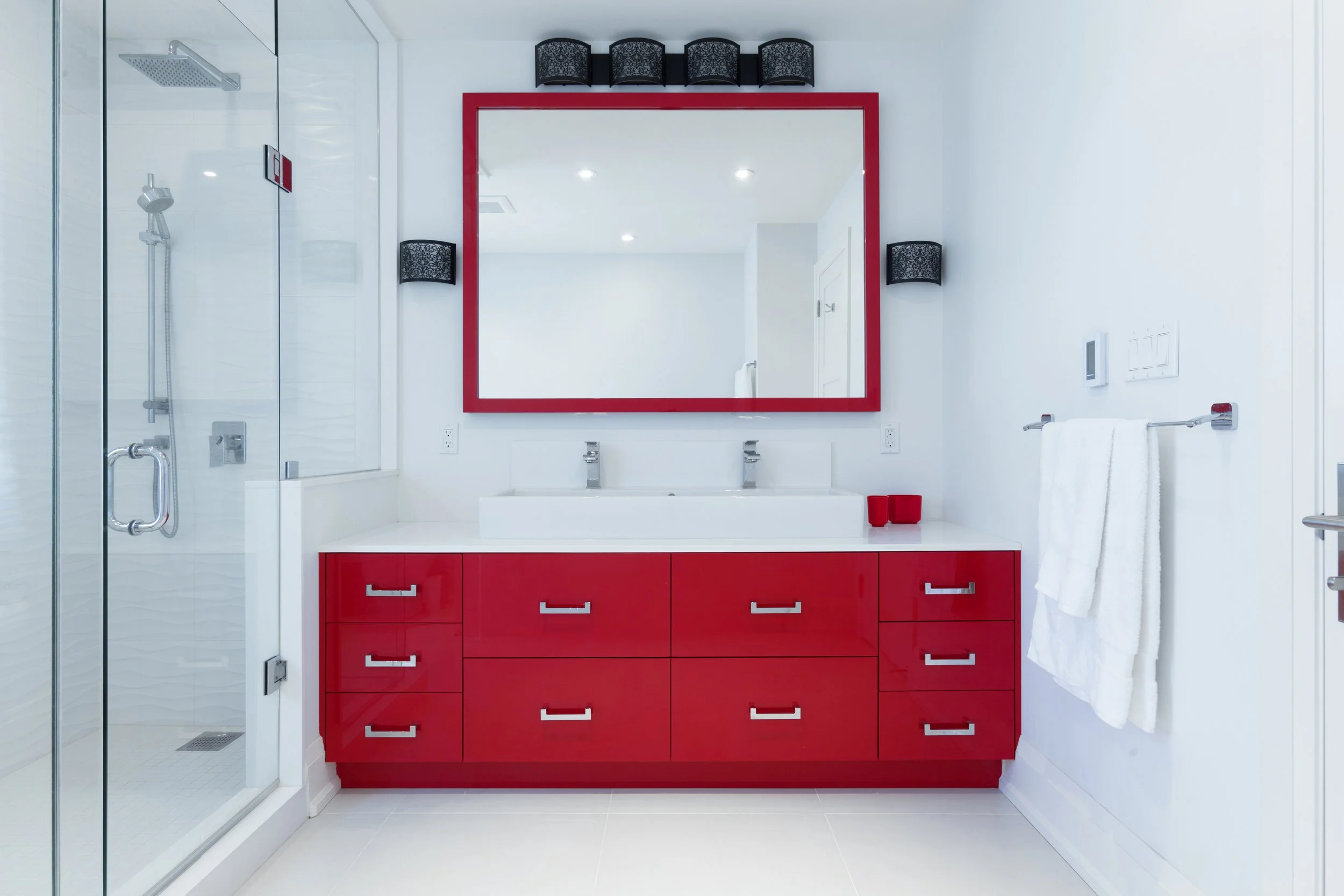 Modern bathroom with a red vanity with multiple drawers, large mirror, black decorative wall lamps, towels on a silver towel bar, and a glass shower enclosure on the left.