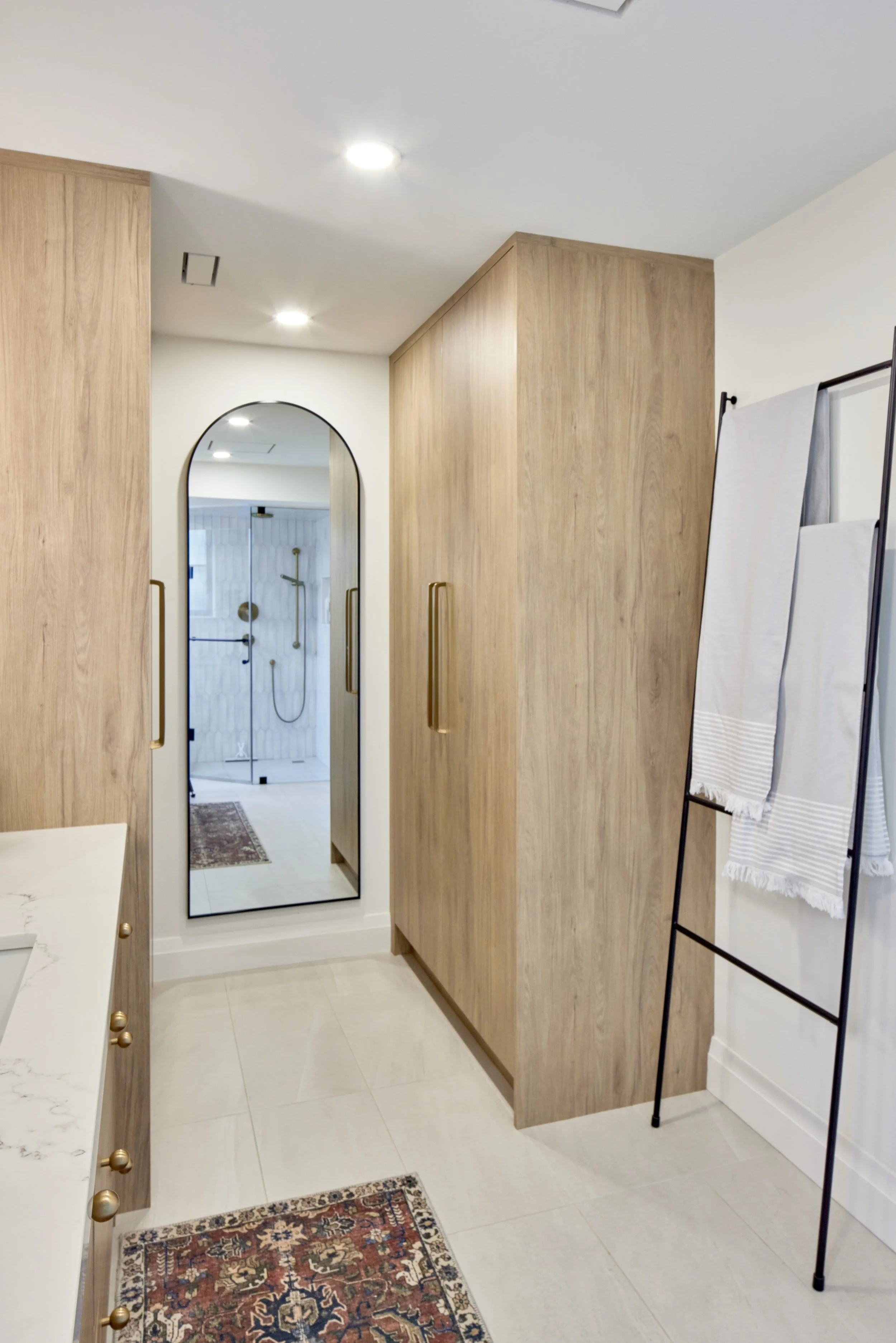 A room with light-colored tile flooring, wooden cabinets, a tall mirror, a decorative rug, and a clothing rack with towels. A door leads to a bathroom with a walk-in shower, a round showerhead, and wooden accents.