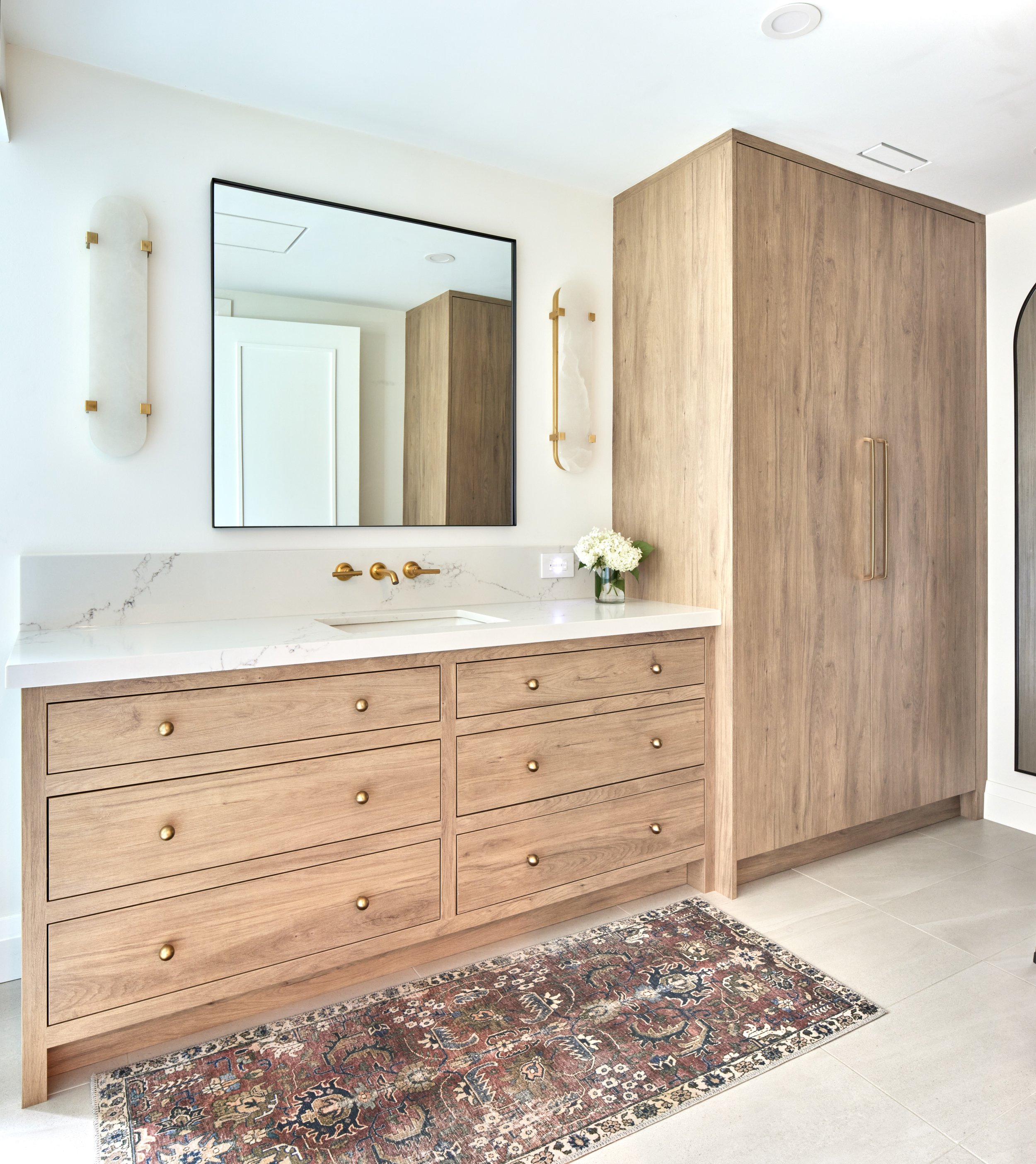 Modern bathroom with light wood vanity and cabinetry, white marble countertop, large mirror, gold fixtures, a small bouquet of white flowers, and a decorative rug on a gray tiled floor.