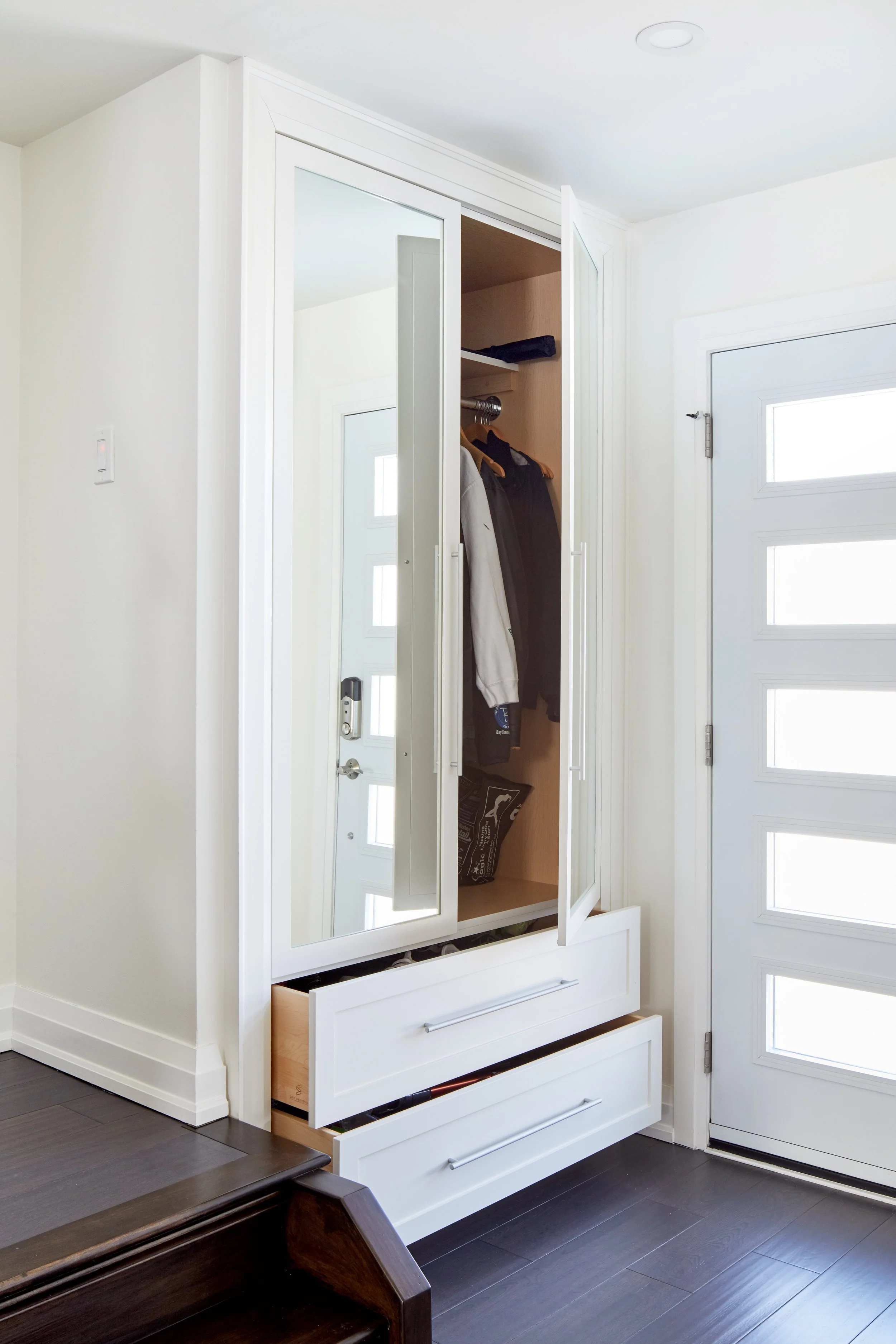 A white wardrobe with a mirror door, open to reveal hanging clothes and a shelf inside, located near a white door with rectangular windows, against white walls and dark flooring.