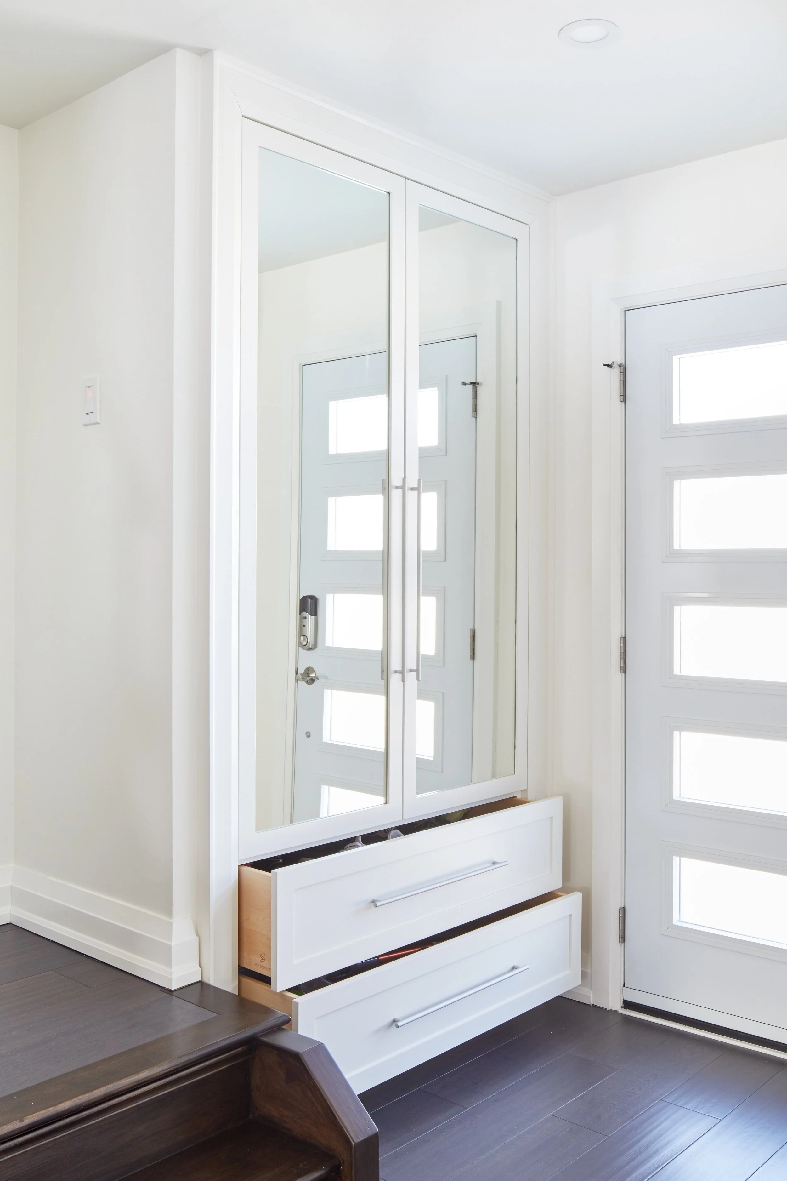 A white built-in closet with mirrored sliding doors in a bright room with white walls, a small horizontal window, dark hardwood flooring, and the bottom drawers of the closet are open, revealing storage space.