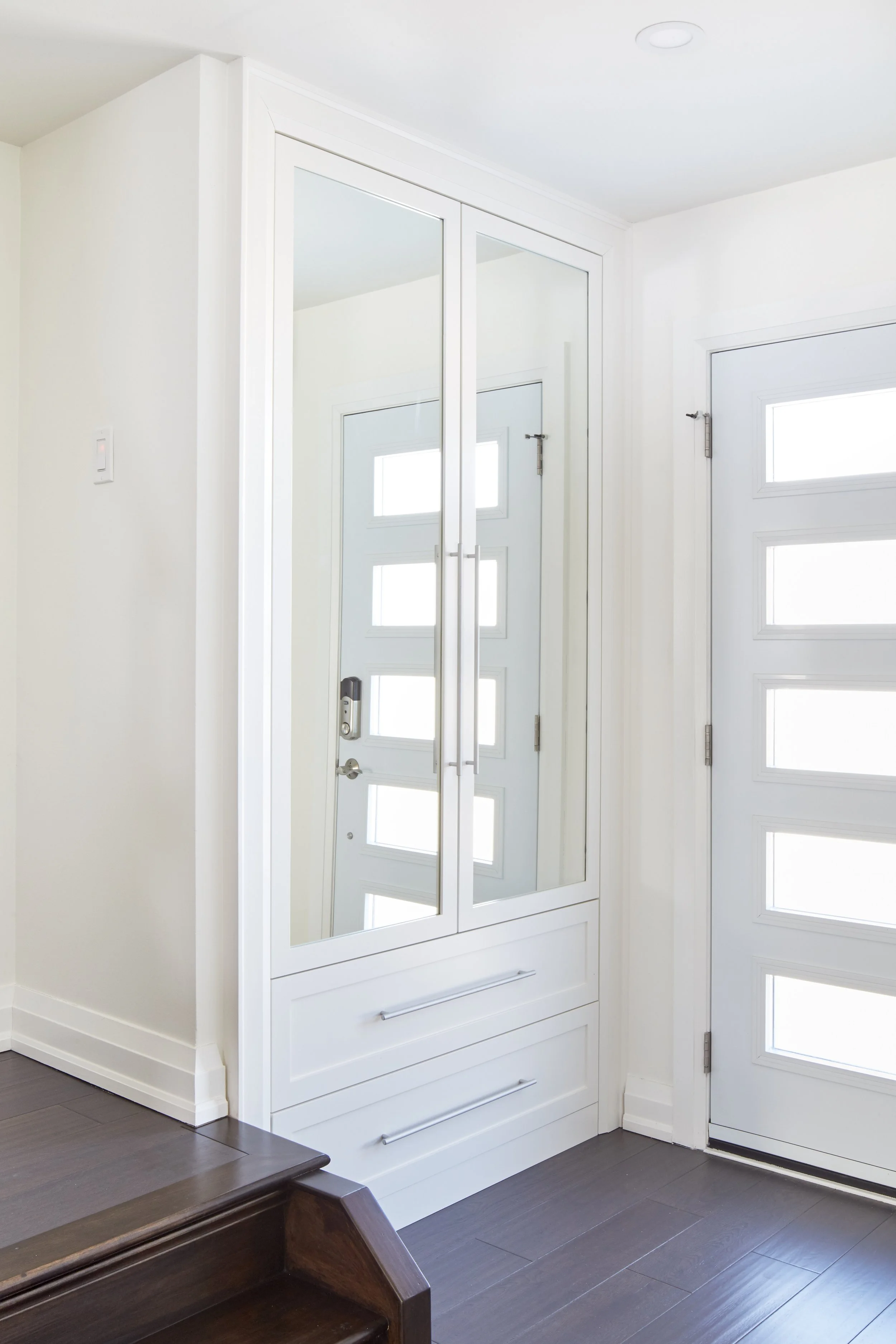 A modern white entryway with a mirrored closet door, a small window on the side, and a dark hardwood floor.