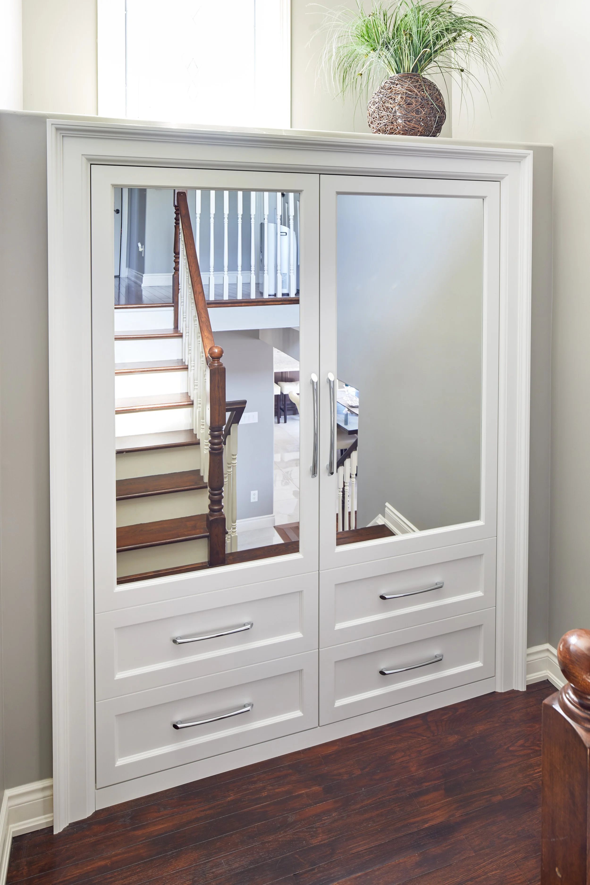 A white wardrobe with sliding mirrors reflecting a staircase and a hallway. The wardrobe has multiple drawers with silver handles and is topped with a decorative woven basket containing a green plant. The room has dark hardwood flooring.