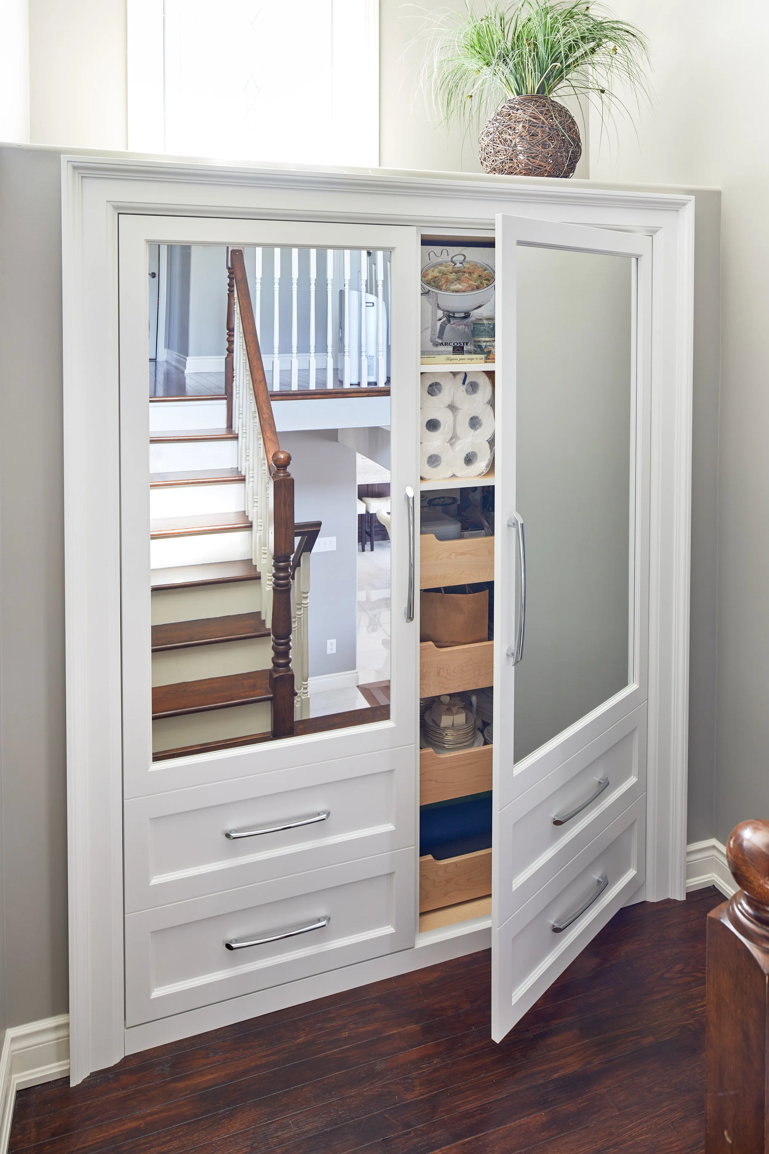 A white cabinet with glass sliding doors reflecting a staircase with wooden steps and white balusters, a potted plant on top, and various household items inside and around.