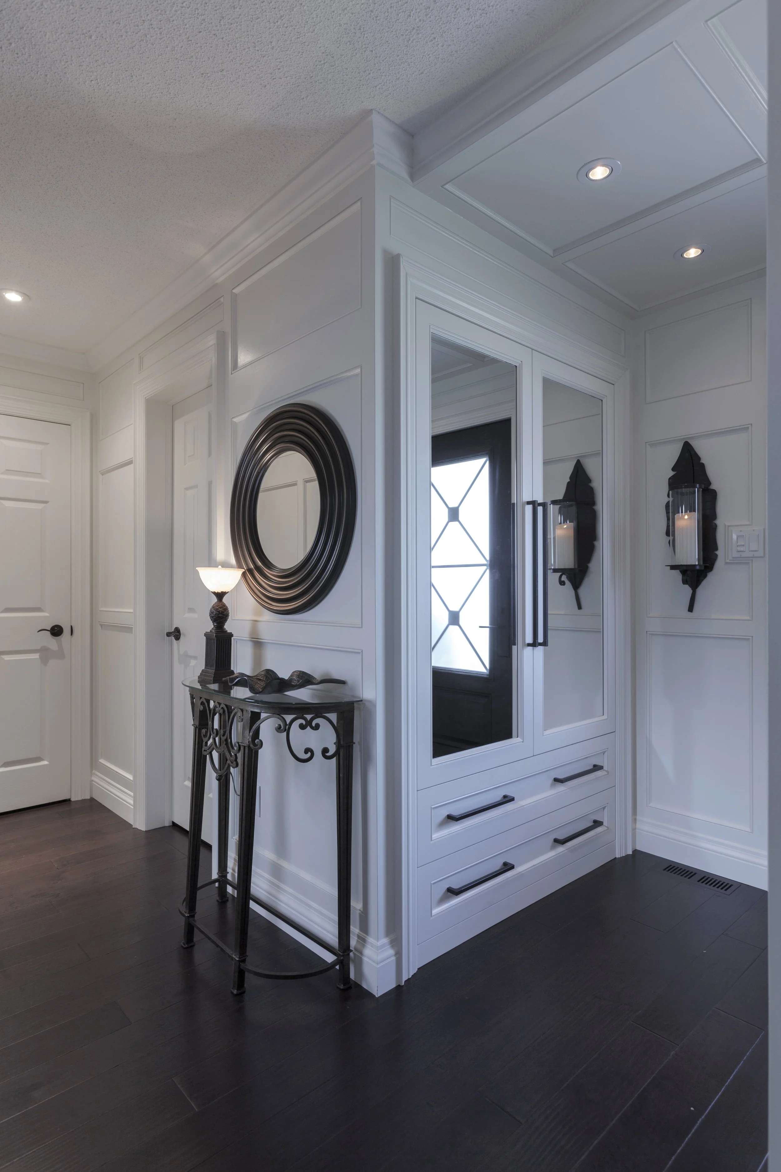 Interior view of a modern entryway with white paneled walls, dark hardwood floors, a built-in closet with glass doors and black handles, and wall-mounted candle sconces. A small black metal table with decorative items stands near the wall. Recessed c