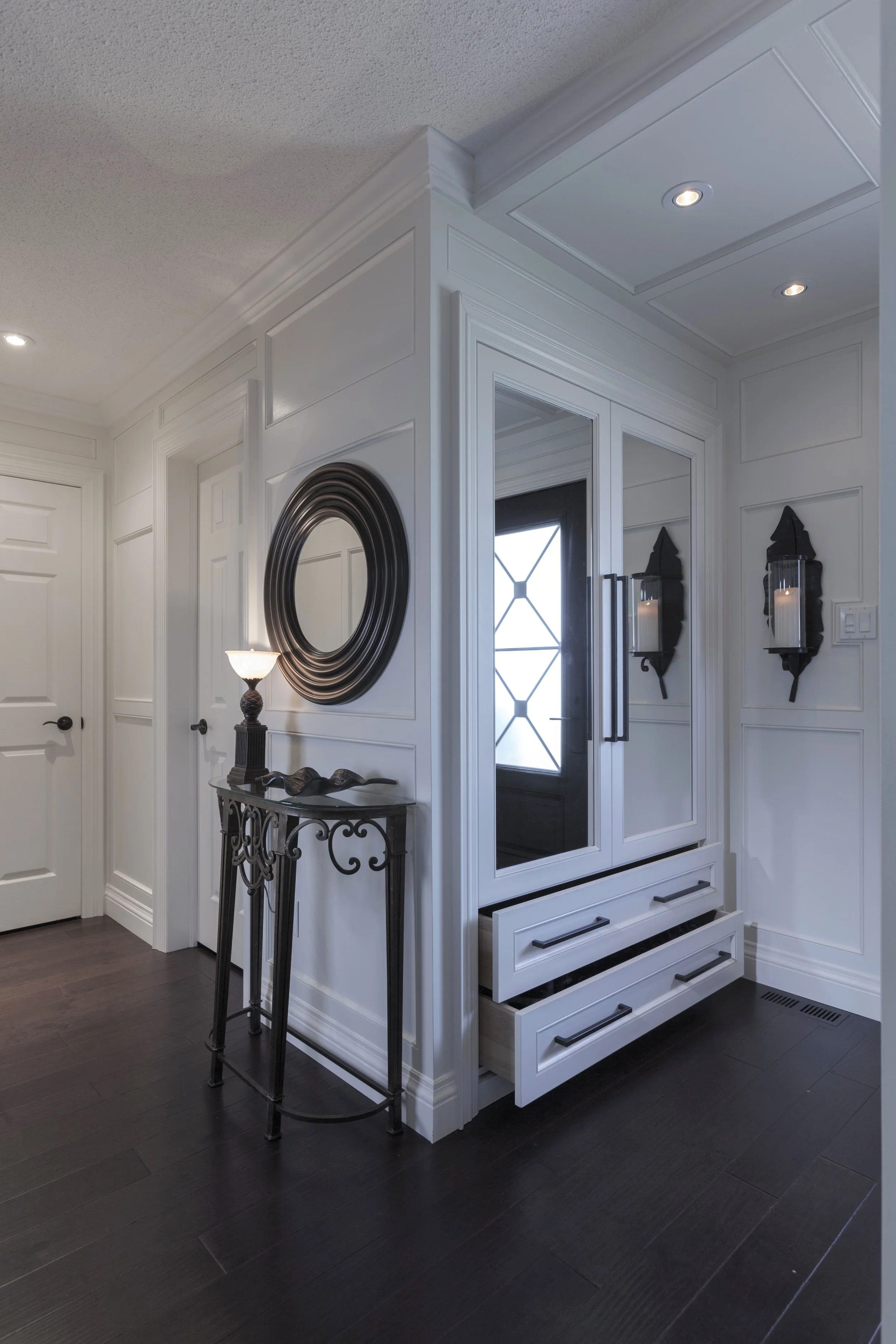 A white entryway with black accents, featuring a large decorative mirror, a black side table with a lamp, and two black wall-mounted candle holders flanking the entrance door, which has glass panels and a geometric design.