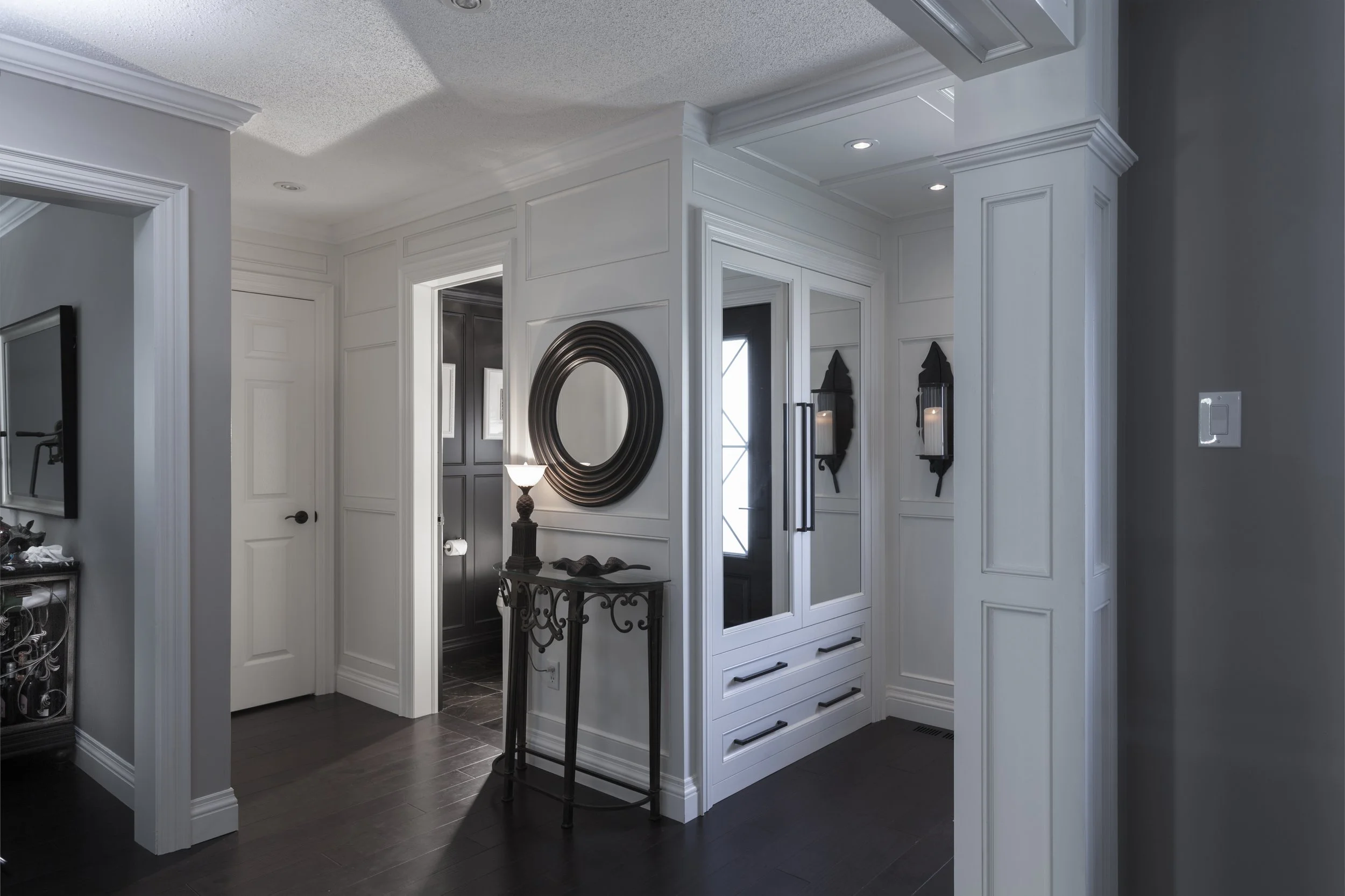Interior view of a modern home entryway with white wall paneling, black accents, a mirror, and dark hardwood flooring.