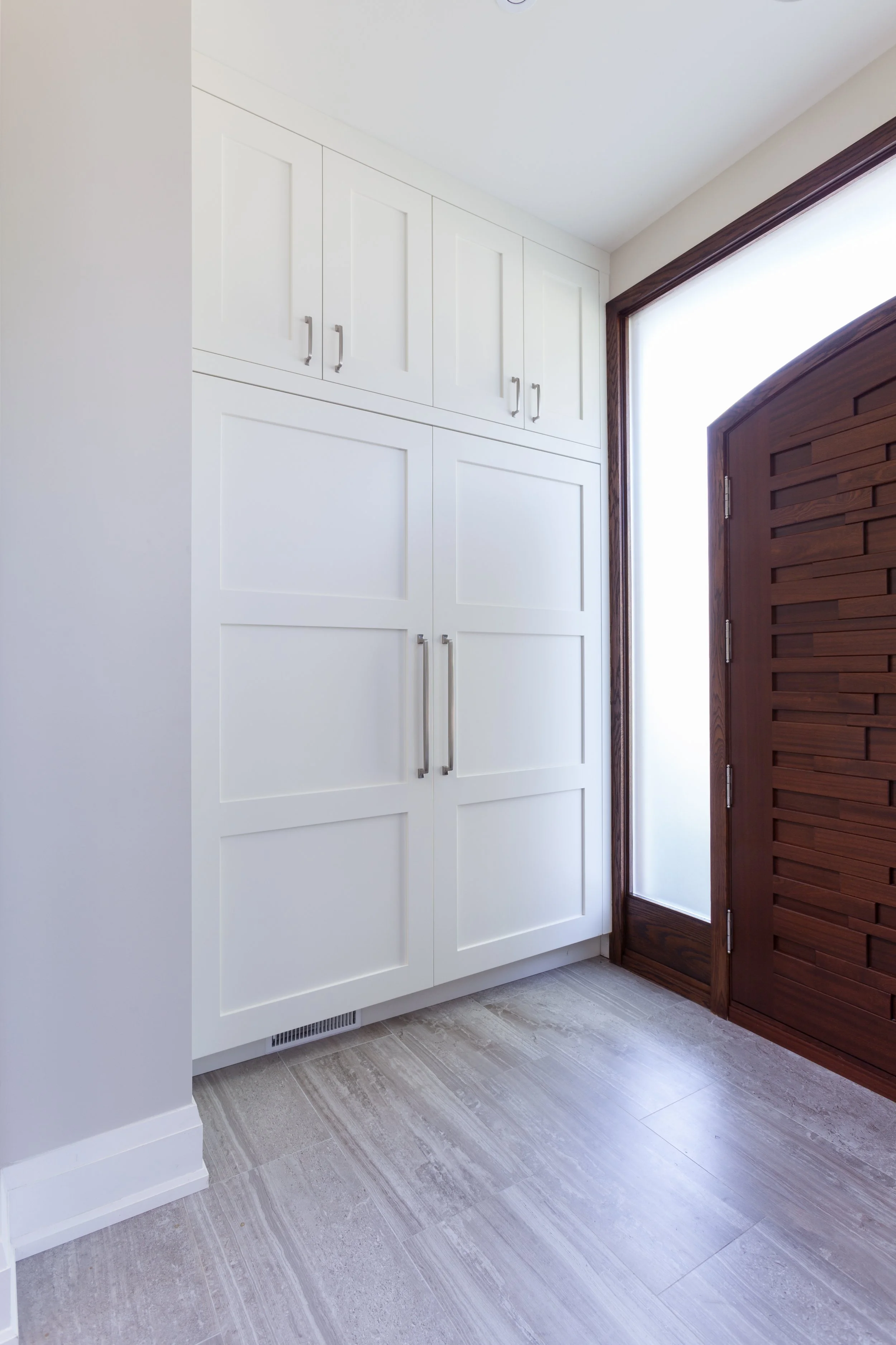Bright entryway with white cabinets, a wooden door with horizontal slats, light-colored tile flooring, and natural light coming through a window.