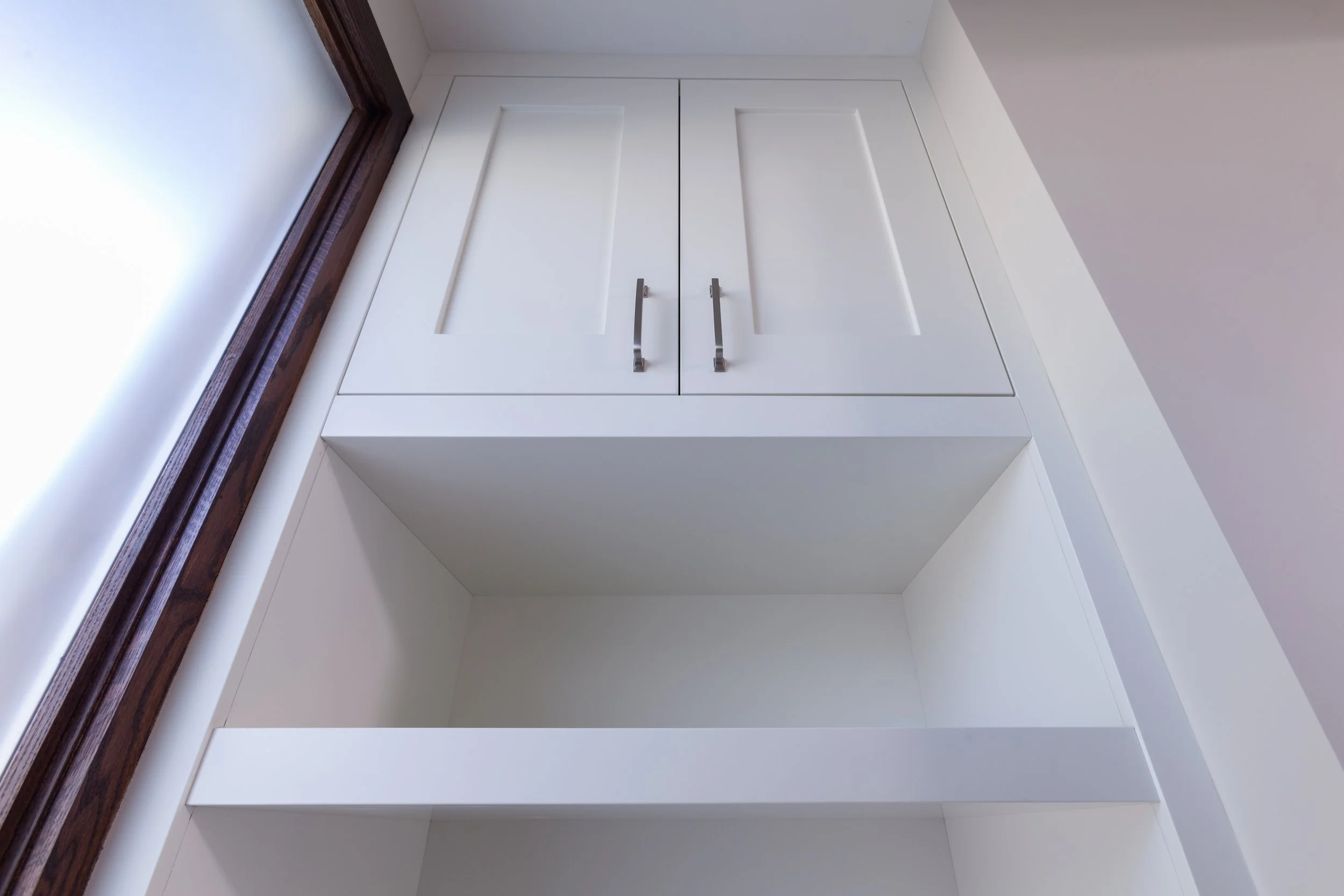 White cabinet with silver handles, built-in shelves underneath, next to a window with wooden trim, in a room with white walls.