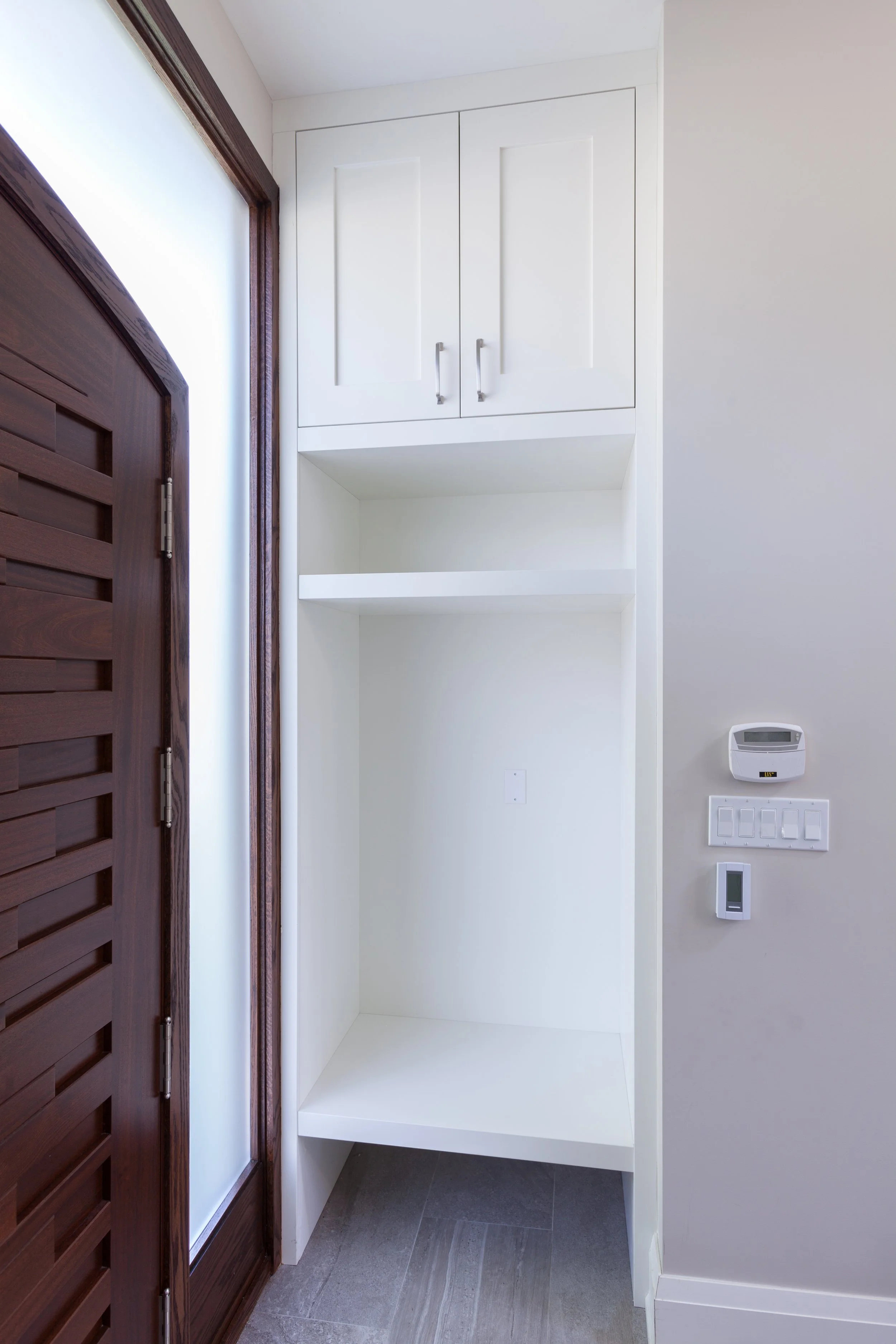 Empty white cabinet with two doors above an open space, next to a brown wooden door with slats, on a wall with a grey tile floor, a thermostat, and light switches.