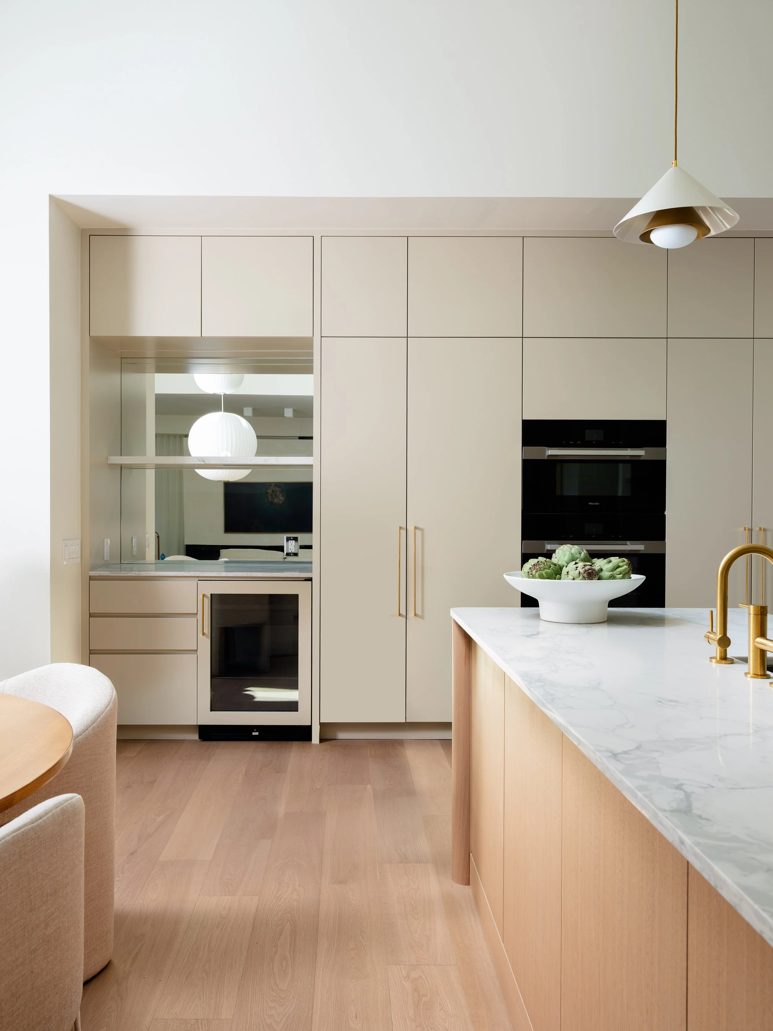 A modern kitchen with built-in cabinets, a marble kitchen island with a gold faucet, a bowl of artichokes, and a large pendant light.