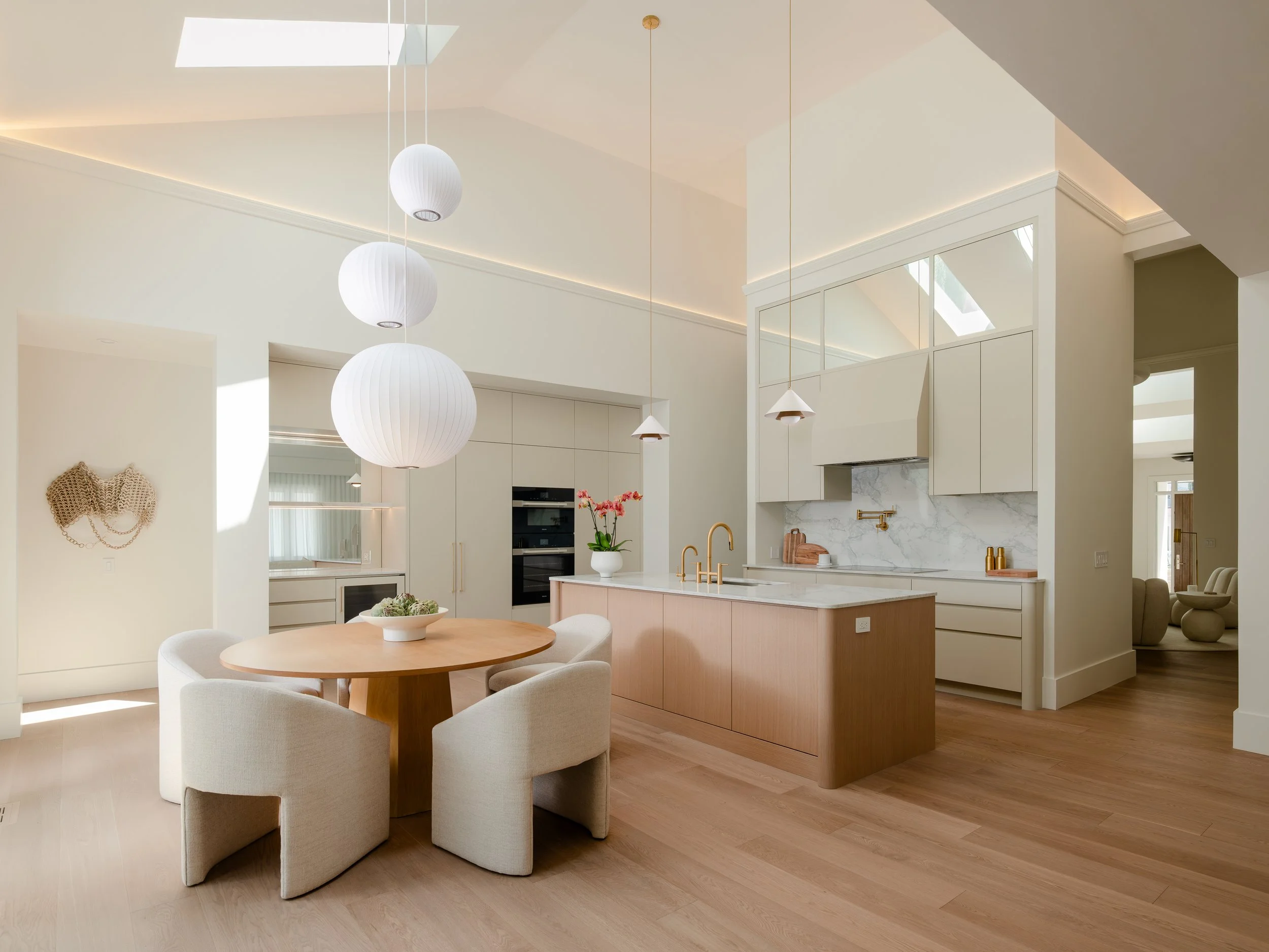 Modern open-concept kitchen and dining area with white walls, natural wood flooring, and minimalist decor. Features white pendant lights, a round wooden dining table with beige chairs, and a kitchen island with a marble backsplash and gold fixtures.