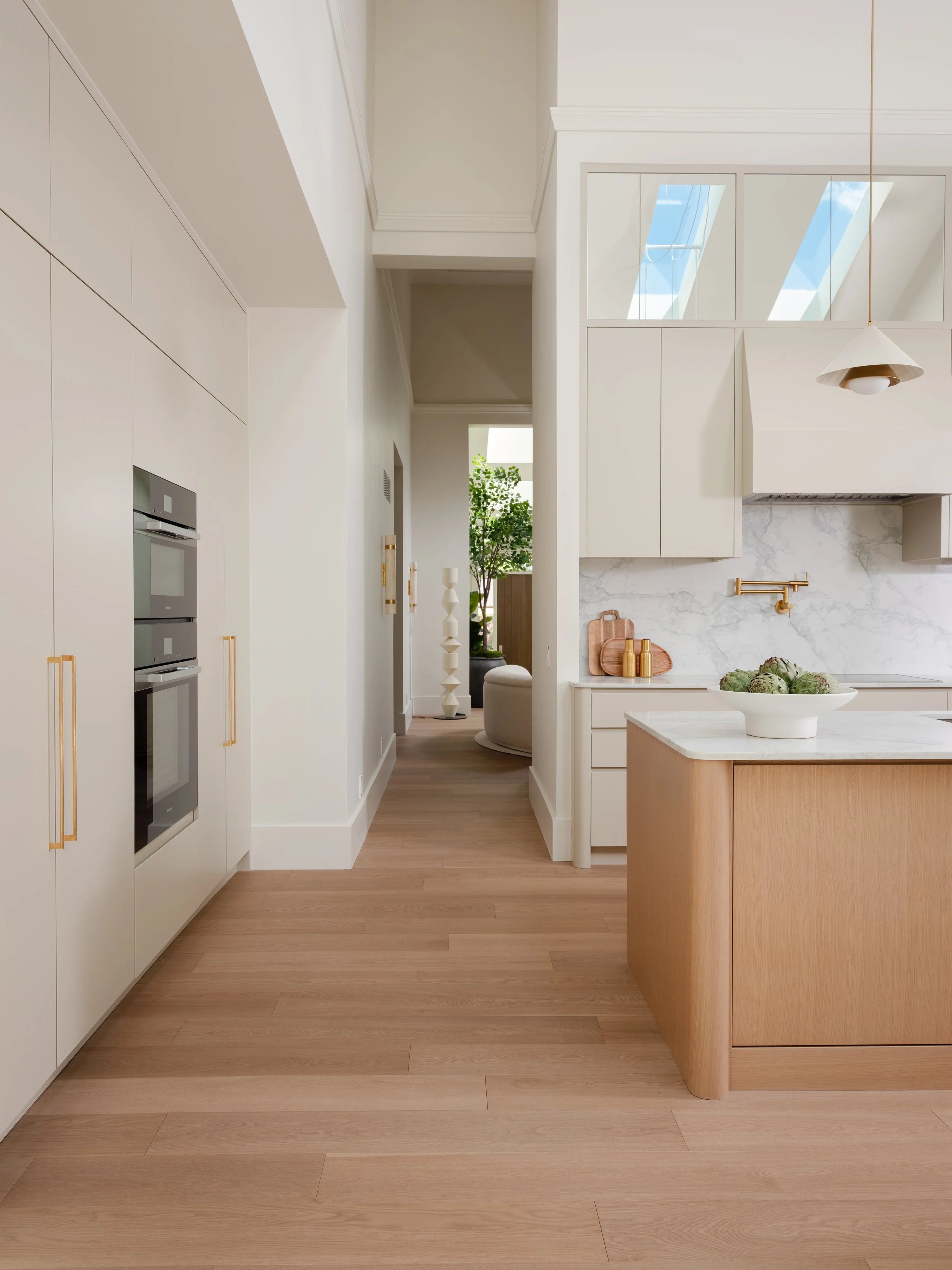 A modern kitchen with light wood flooring, white cabinetry, a marble backsplash, and a large kitchen island with a bowl of artichokes on it.